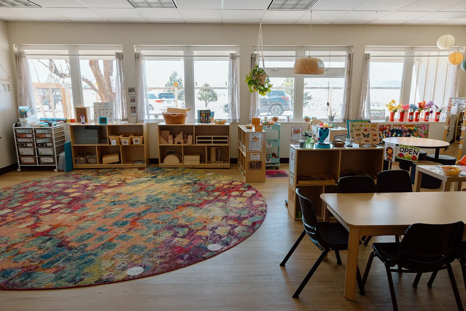 Bright classroom with large windows, colorful circular rug, shelves with educational materials, round table with chairs, and decorations including flowers in vases, paper lanterns, and signs.