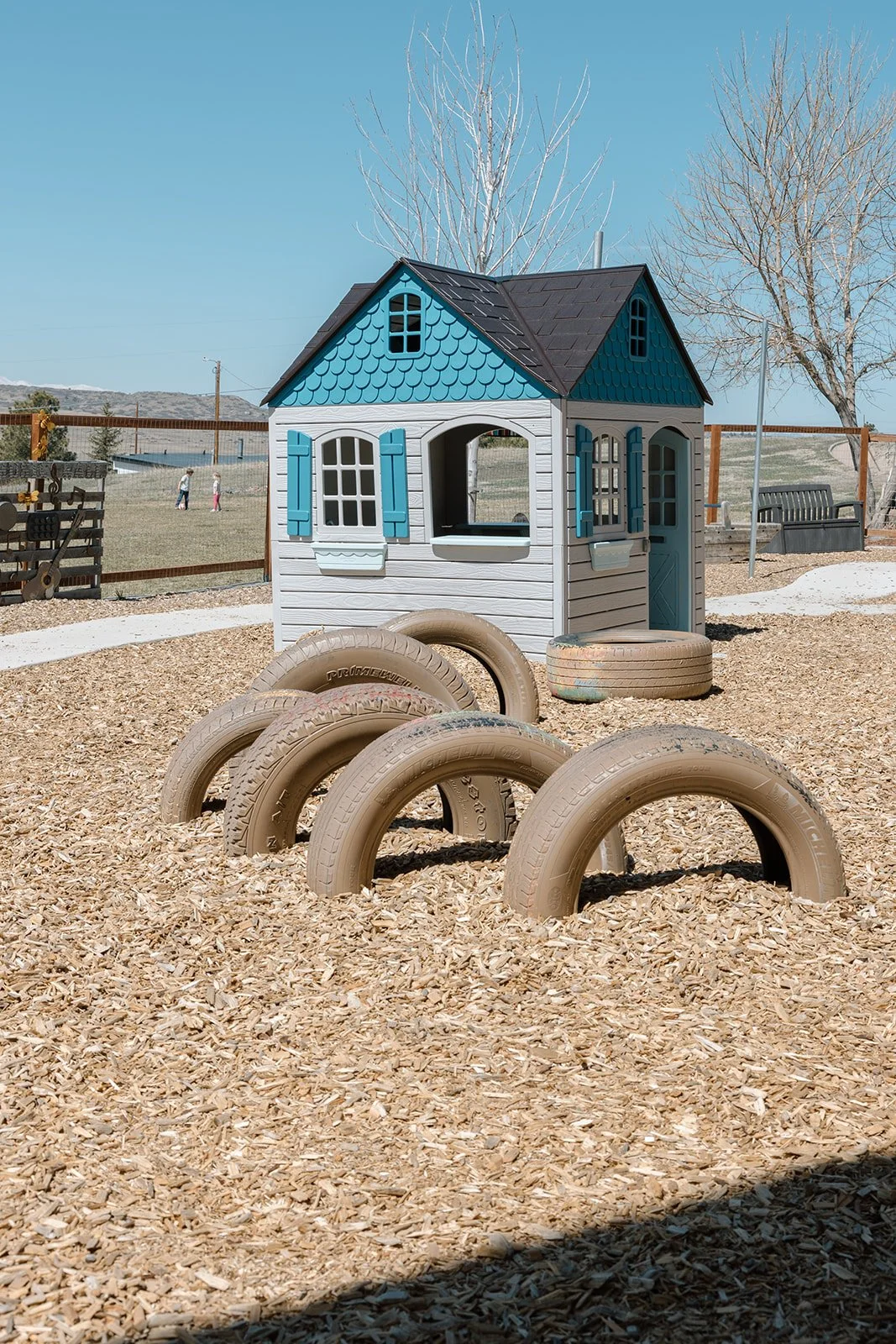 A colorful playhouse with a blue roof and white walls, surrounded by tan tires partially buried in wood chips at a playground. In the background, children are seen playing and a clear blue sky is overhead.