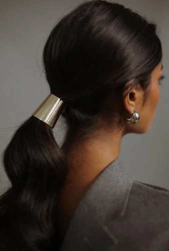 Back of woman’s head with dark hair styled in a ponytail secured with a wide gold hair tie, wearing a gray blazer and silver hoop earrings.
