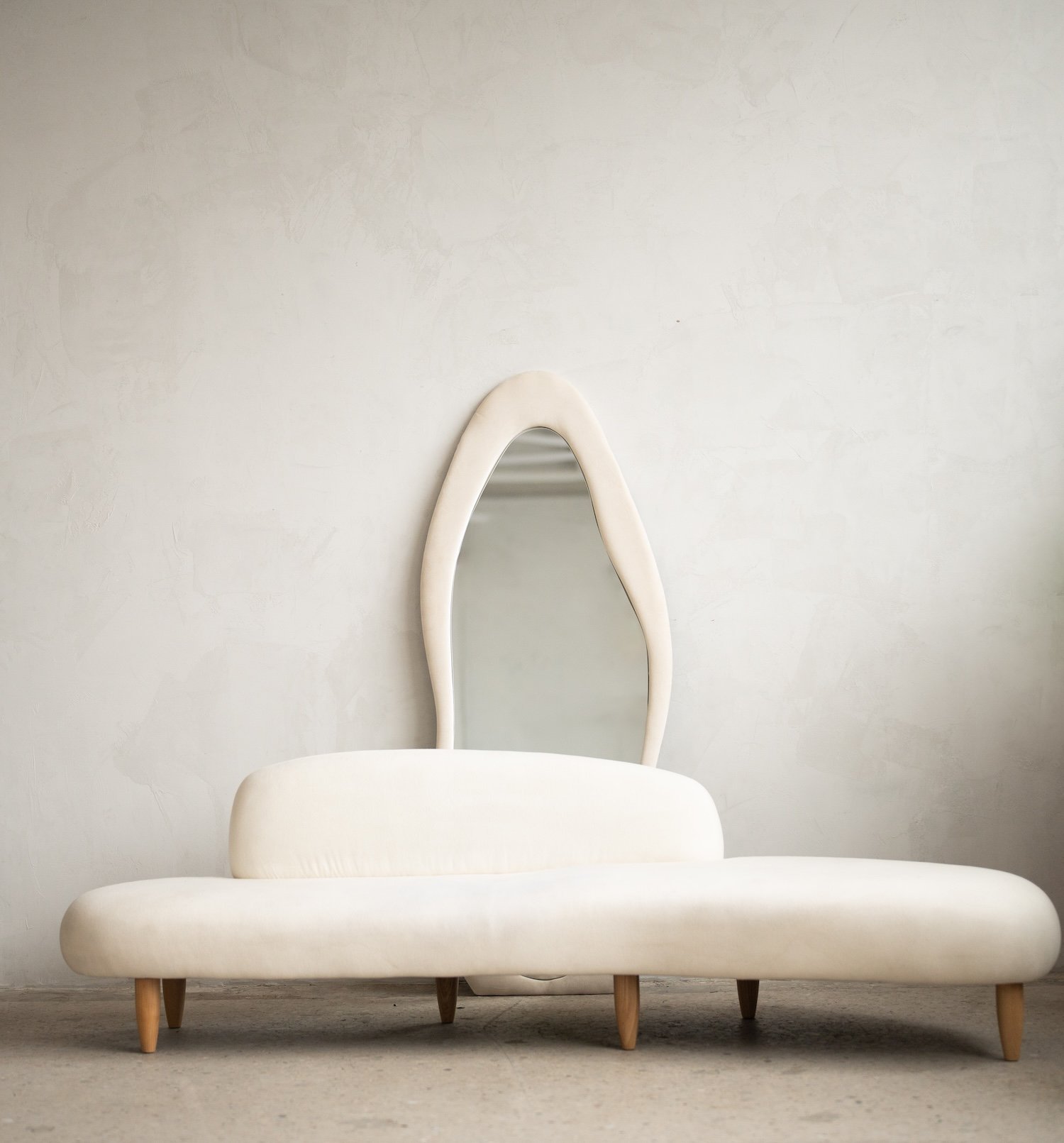 White modern furniture with a large cushion and a tall, irregular-shaped mirror behind it in a minimalist room.