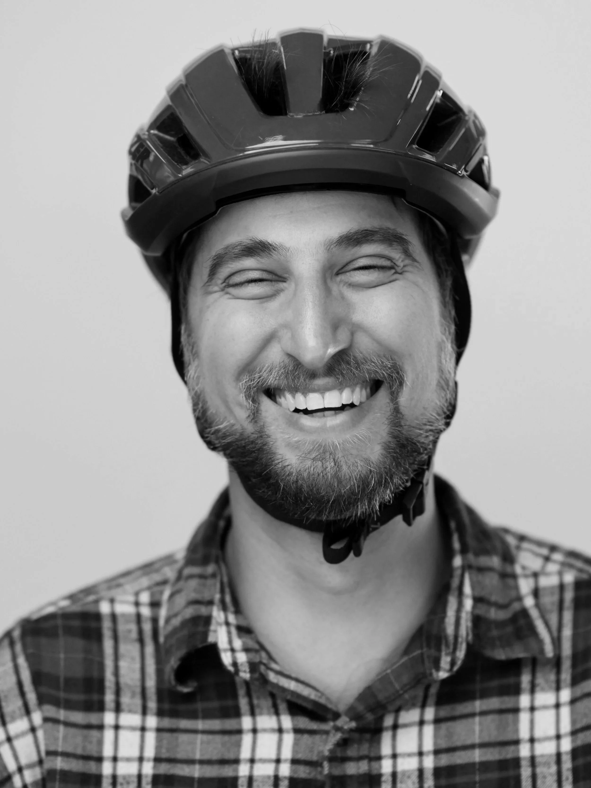 A smiling man wearing a red bicycle helmet and a plaid shirt.