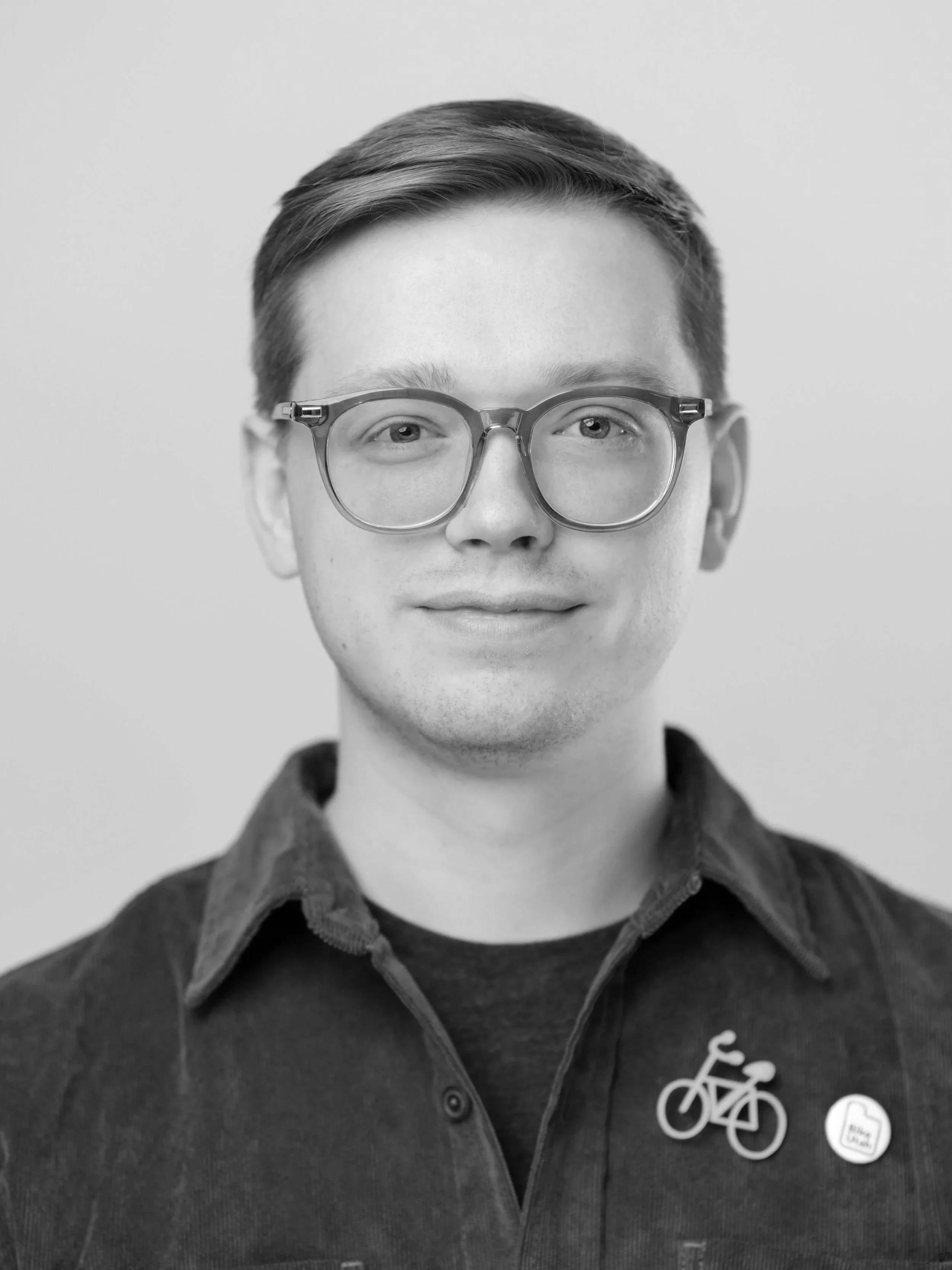 A young man with light skin, glasses, and reddish-brown hair smiling at the camera. He is wearing a green jacket with a bicycle pin and a speech bubble pin.