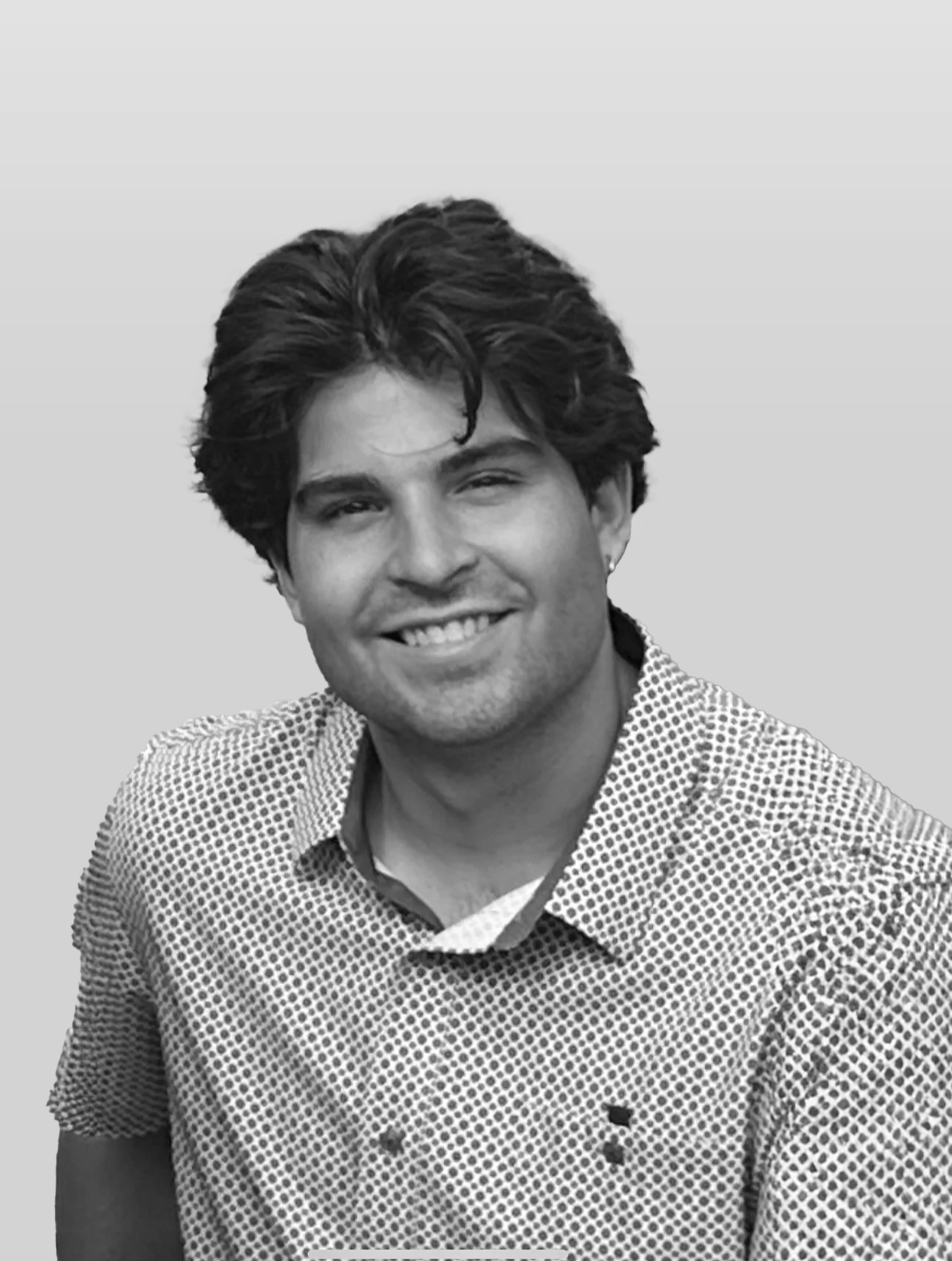 A young man with dark, wavy hair and a light beard, smiling and wearing a patterned button-up shirt, standing against a plain background.