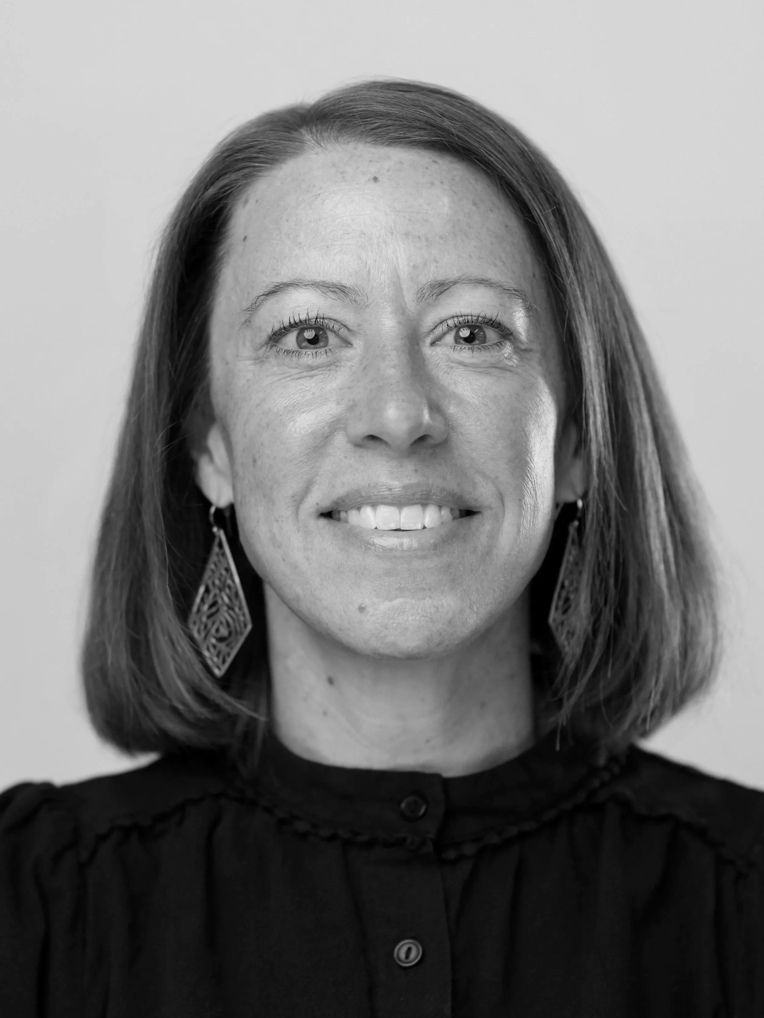 A smiling woman with shoulder-length brown hair, wearing gold earrings and a black buttoned blouse, posing against a plain light gray background.