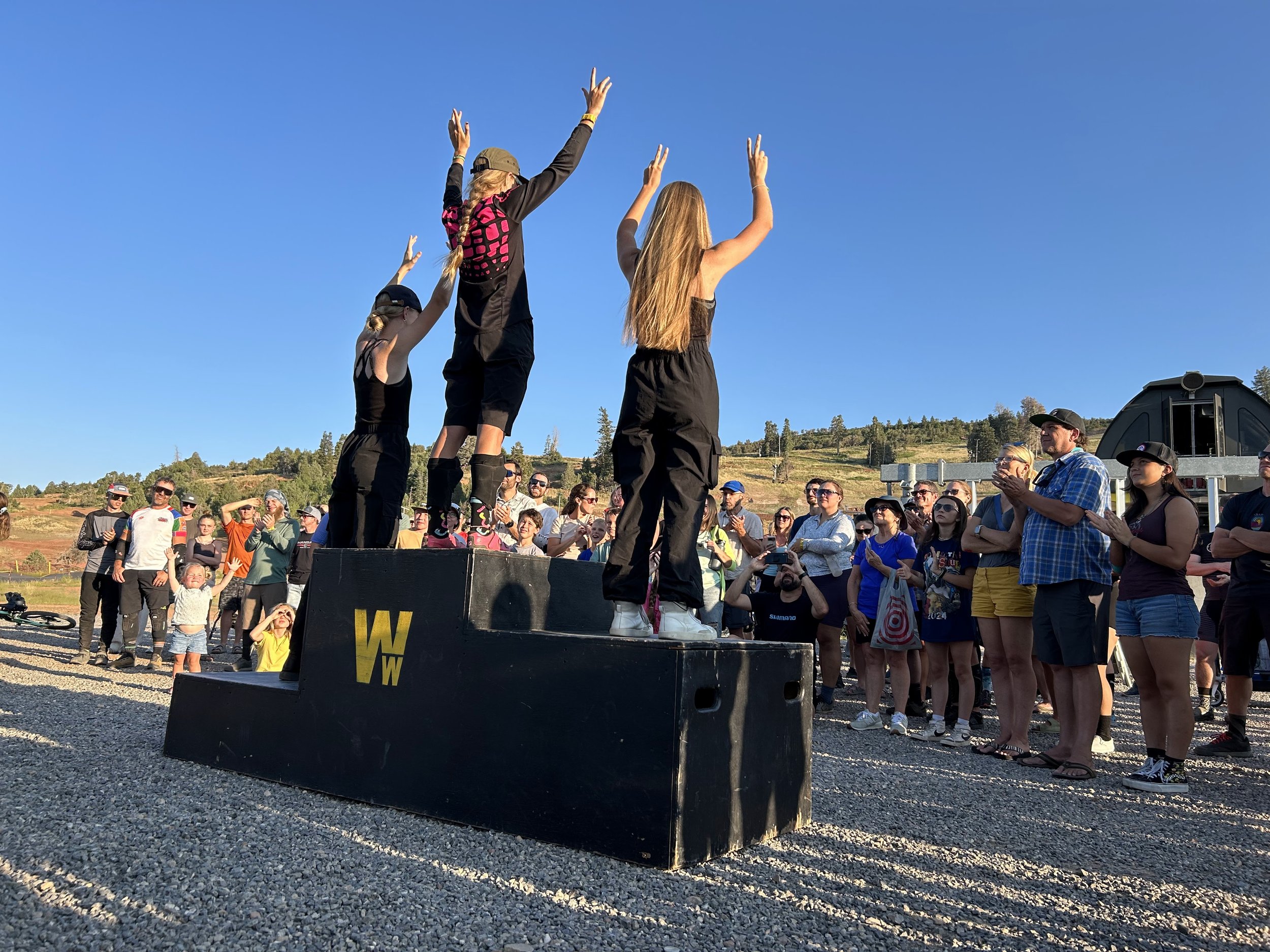 Three women atop a podium with a crowd in front of them