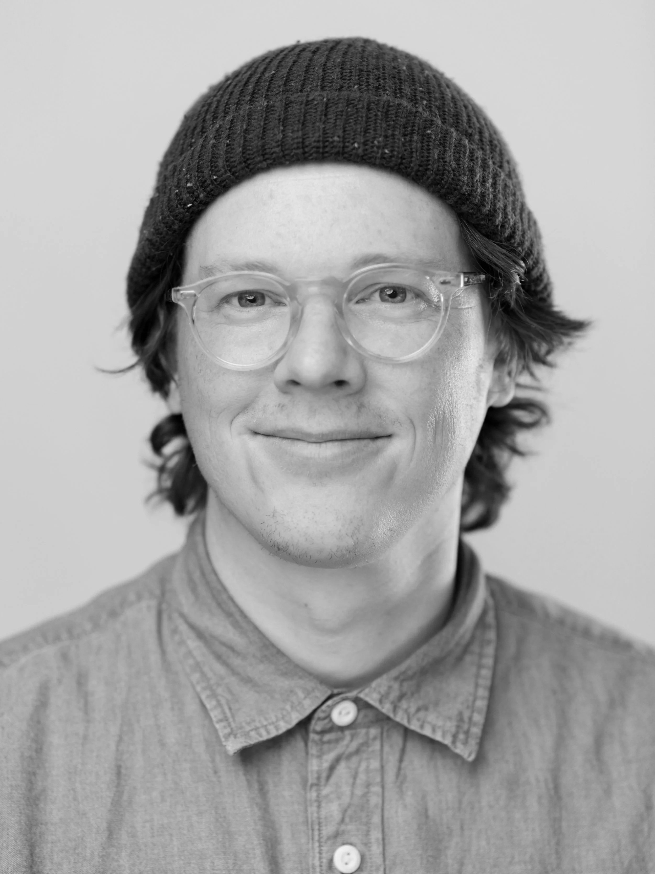 Young man with light skin, brown hair, glasses, and a beanie, smiling, wearing a blue button-up shirt.
