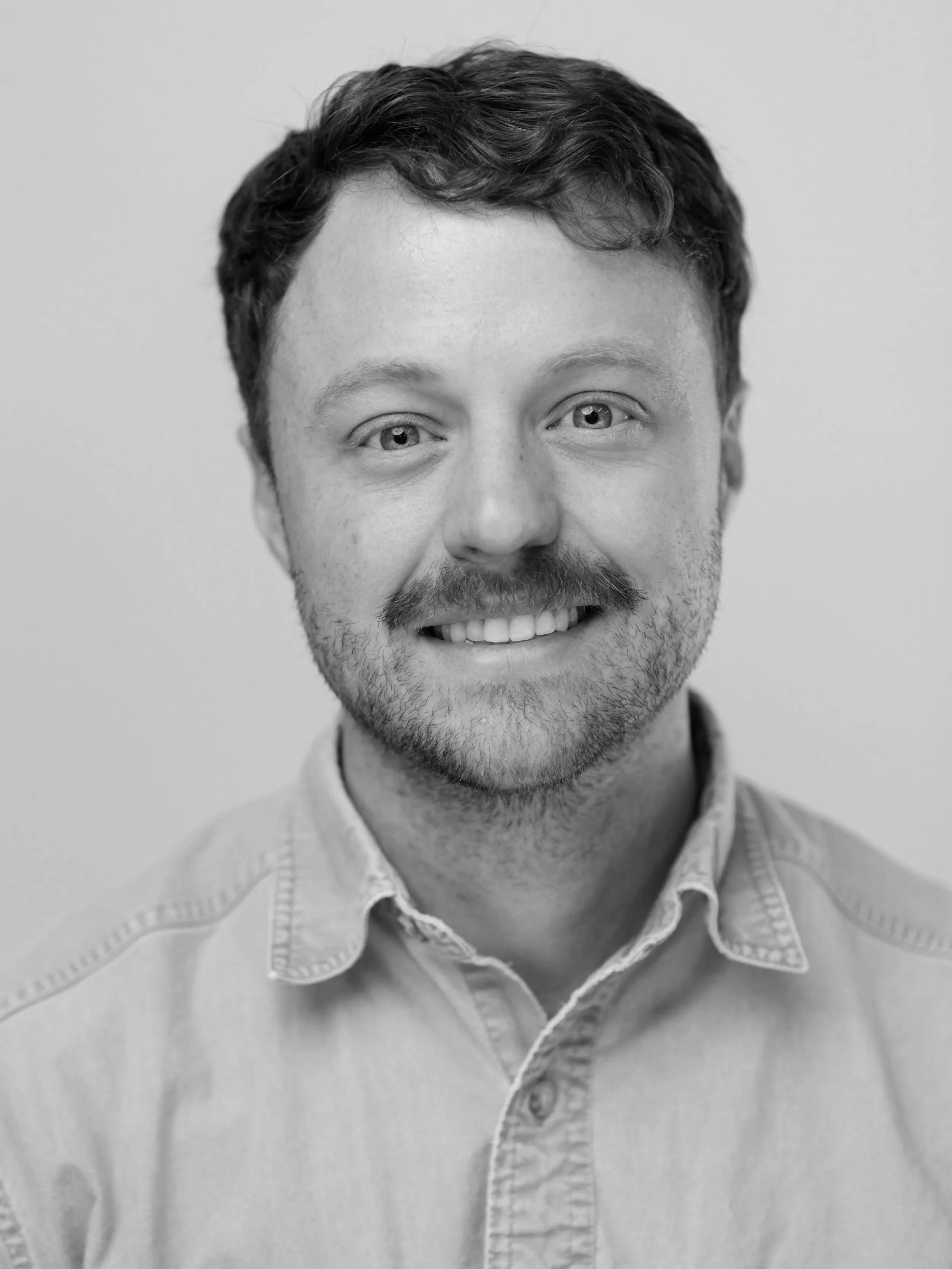 Close-up portrait of a smiling man with blue eyes, brown hair, and a beard, wearing a light blue collared shirt