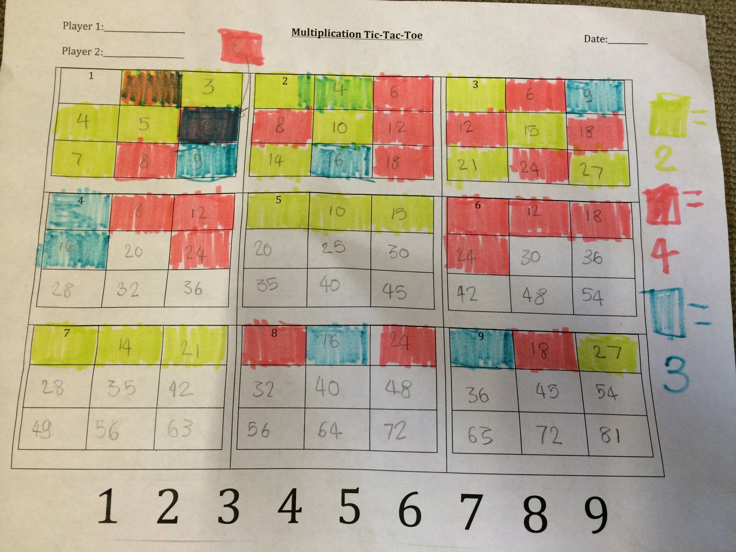 A student's paper multiplication tic-tac-toe board, partly colored in by hand. Caption: A student's own color-coding, found on the floor by the cubbies.