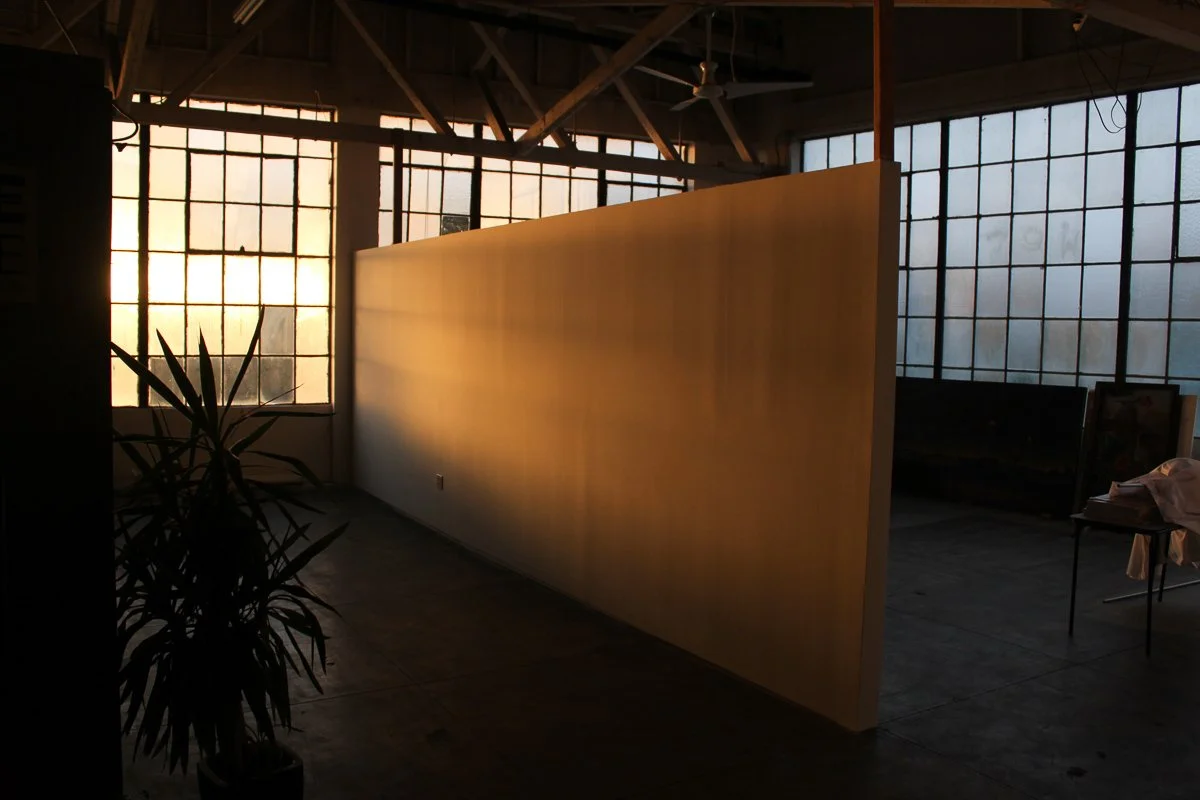 Interior of an industrial-style art studio with large windows, a tall yellow partition, and a potted plant near the left side during sunset.