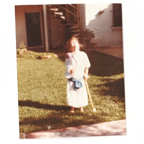 A young girl dressed in a medieval costume standing outdoors on a grassy area holding a wooden stick, with a house and garden in the background.
