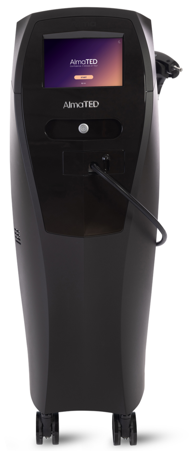 A black medical device with a touchscreen display showing the AlmaTED logo and the words "Confidence. It Grows on You!" in purple and white, with a start button. The device has a circular button below the screen, a flexible hose attached to the right side, and is mounted on four wheels for mobility.