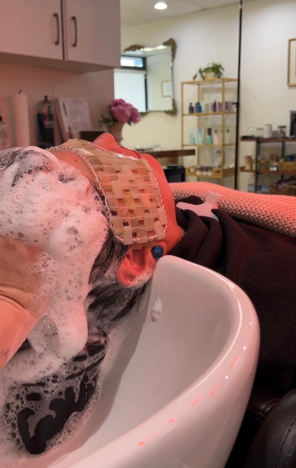 Person getting a scalp treatment in a room with a mirror, flowers, and shelves with bottles.