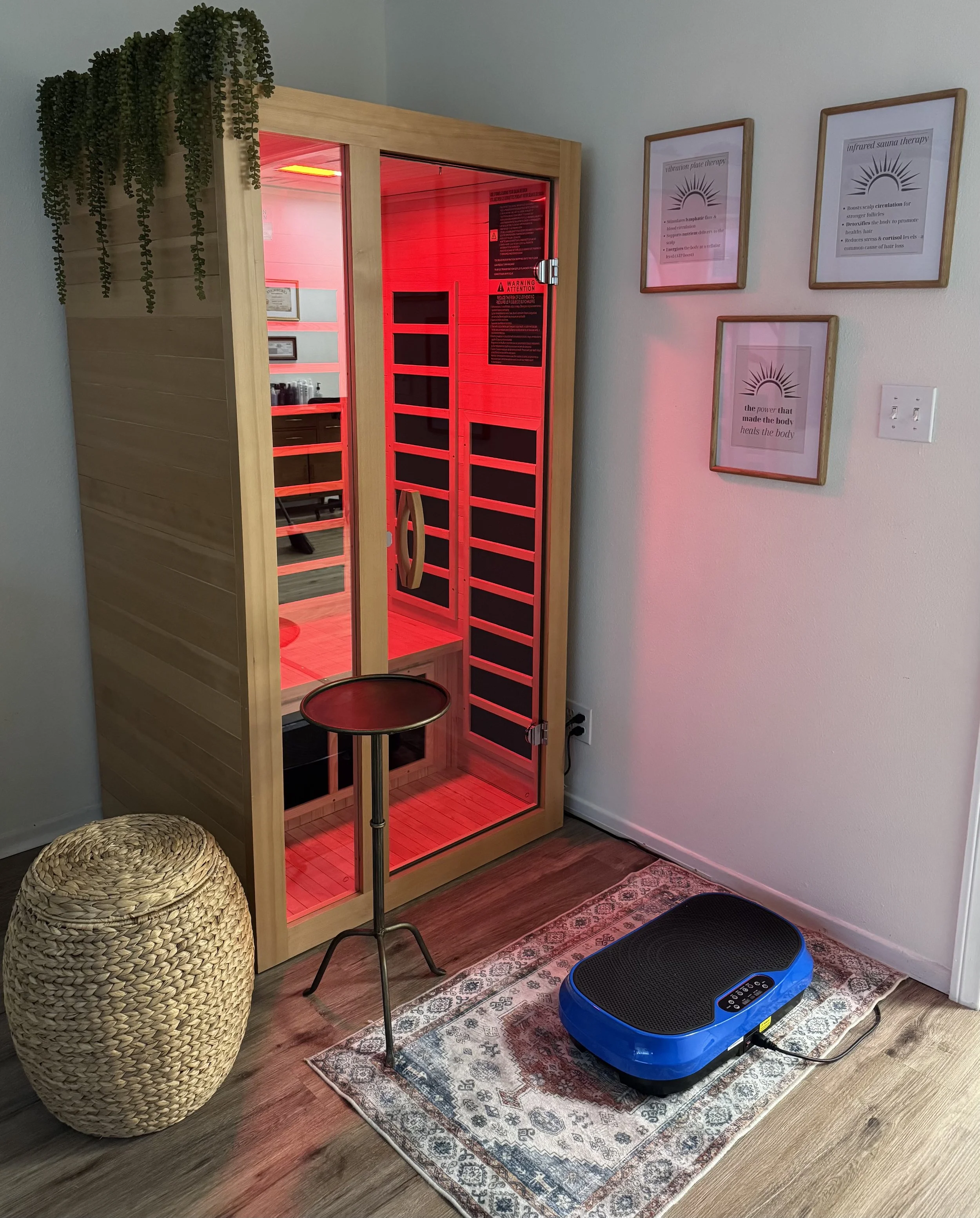A wooden infrared sauna with glass door, red interior lighting, and black control panel, situated in a room with hardwood flooring, area rug, a black/blue vibration plate, a woven wicker basket, and framed informational posters on the wall.