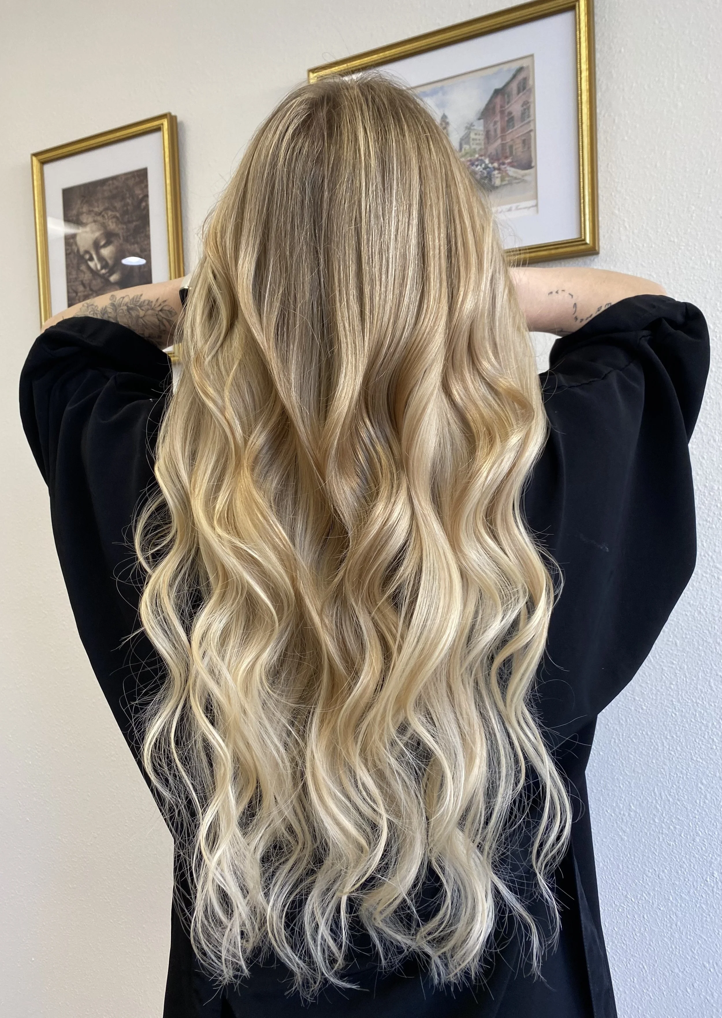 A woman with long, wavy blonde hair stands with her back to the camera, in front of a wall decorated with framed artwork.