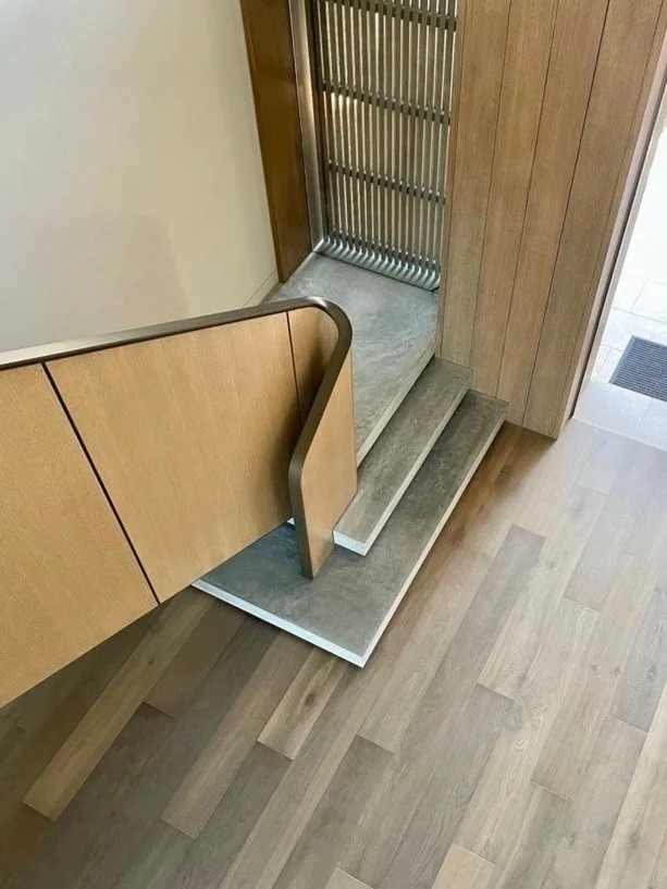 A small set of concrete steps with two steps, leading up to a door with a metal gate, next to a wooden cabinet and light wood flooring.