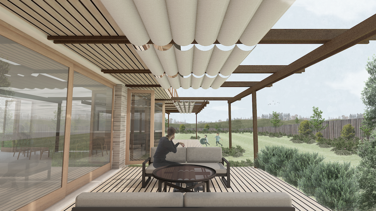 A covered outdoor patio with wooden decking, glass doors, and a person sitting on a sofa, looking at children playing on a grassy yard.