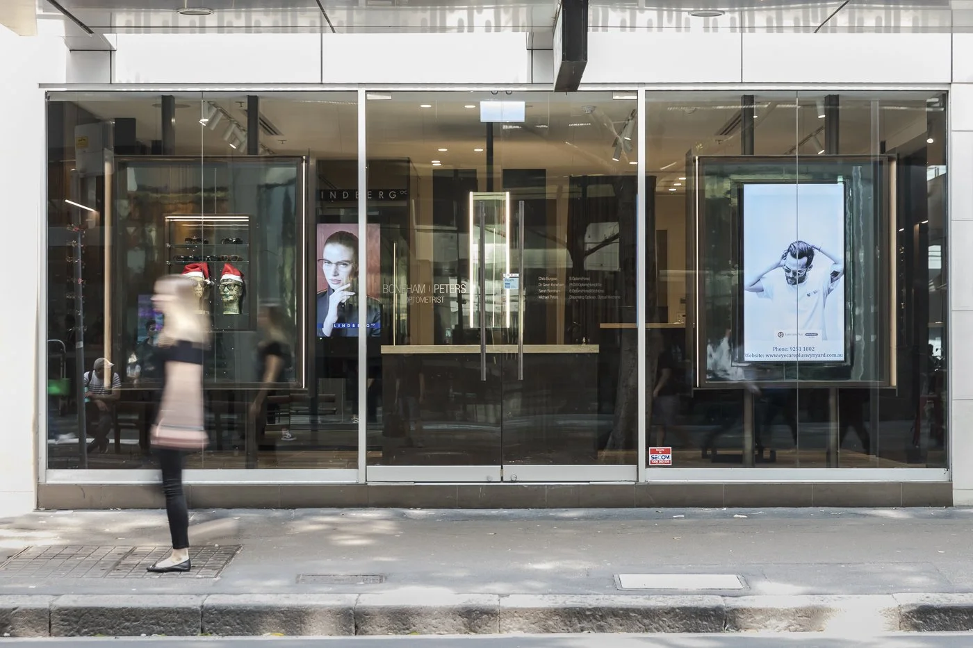 Storefront with glass windows displaying advertisements and optical store signage, with blurred pedestrians walking by on the sidewalk.