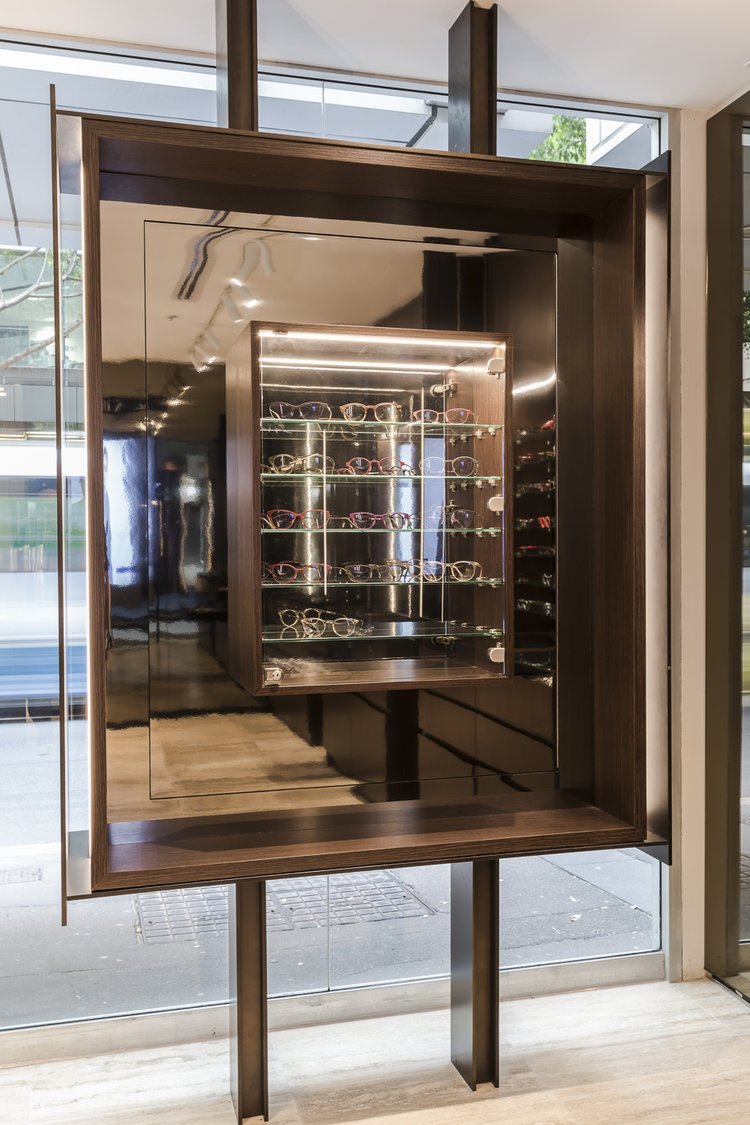 A display case with multiple shelves filled with sunglasses, situated on a wooden stand in a modern store with large glass windows.