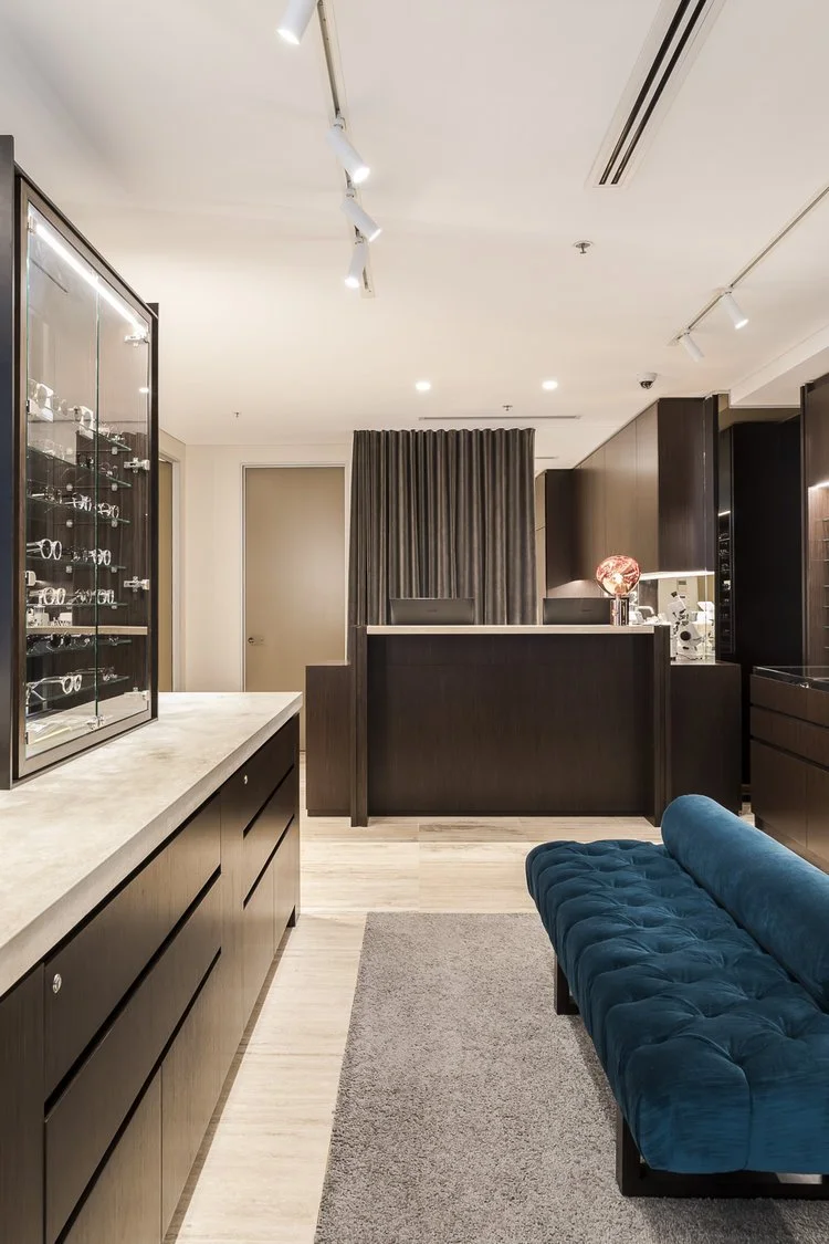 Interior of a modern retail store or showroom with display cases showcasing eyewear, a velvet blue sofa, a beige rug, and a reception desk with decorative items, illuminated by track lighting.