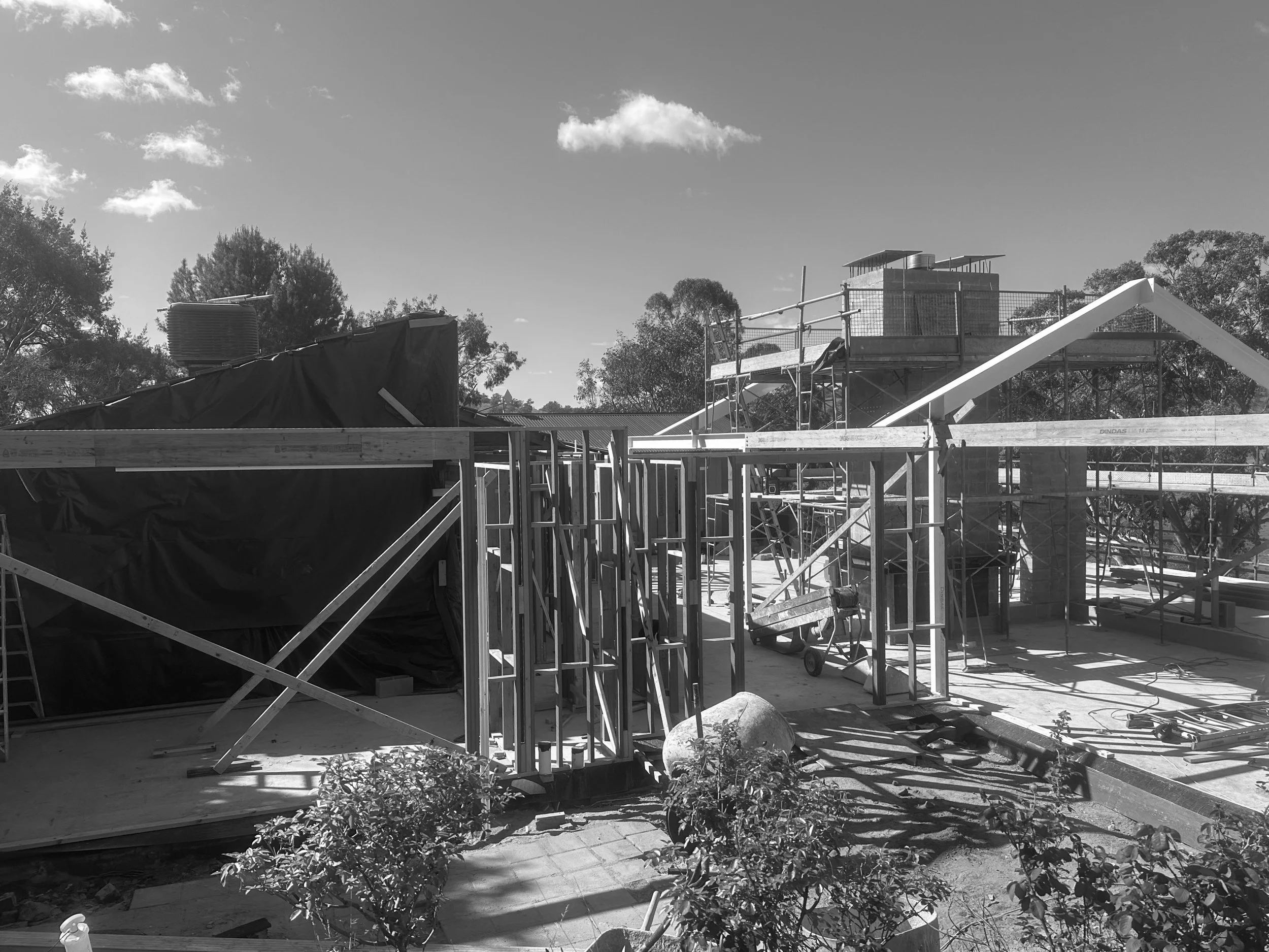 Construction site with wooden framework and scaffolding, residential roofing under construction, trees and sky in the background.