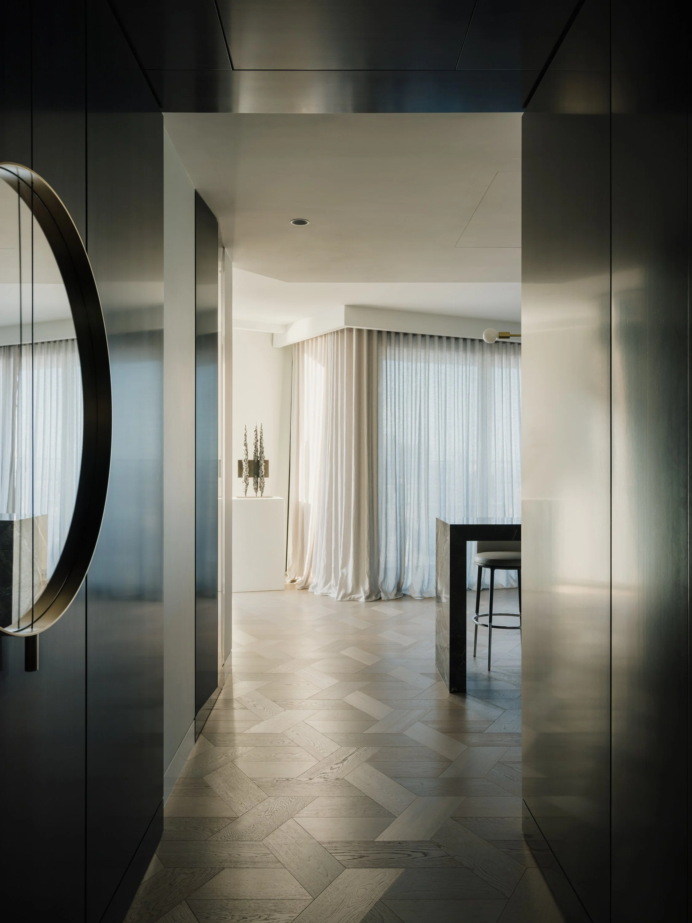 Modern hotel room with floor-to-ceiling curtains, a white pedestal with decorative art, and a barstool at a marble counter.