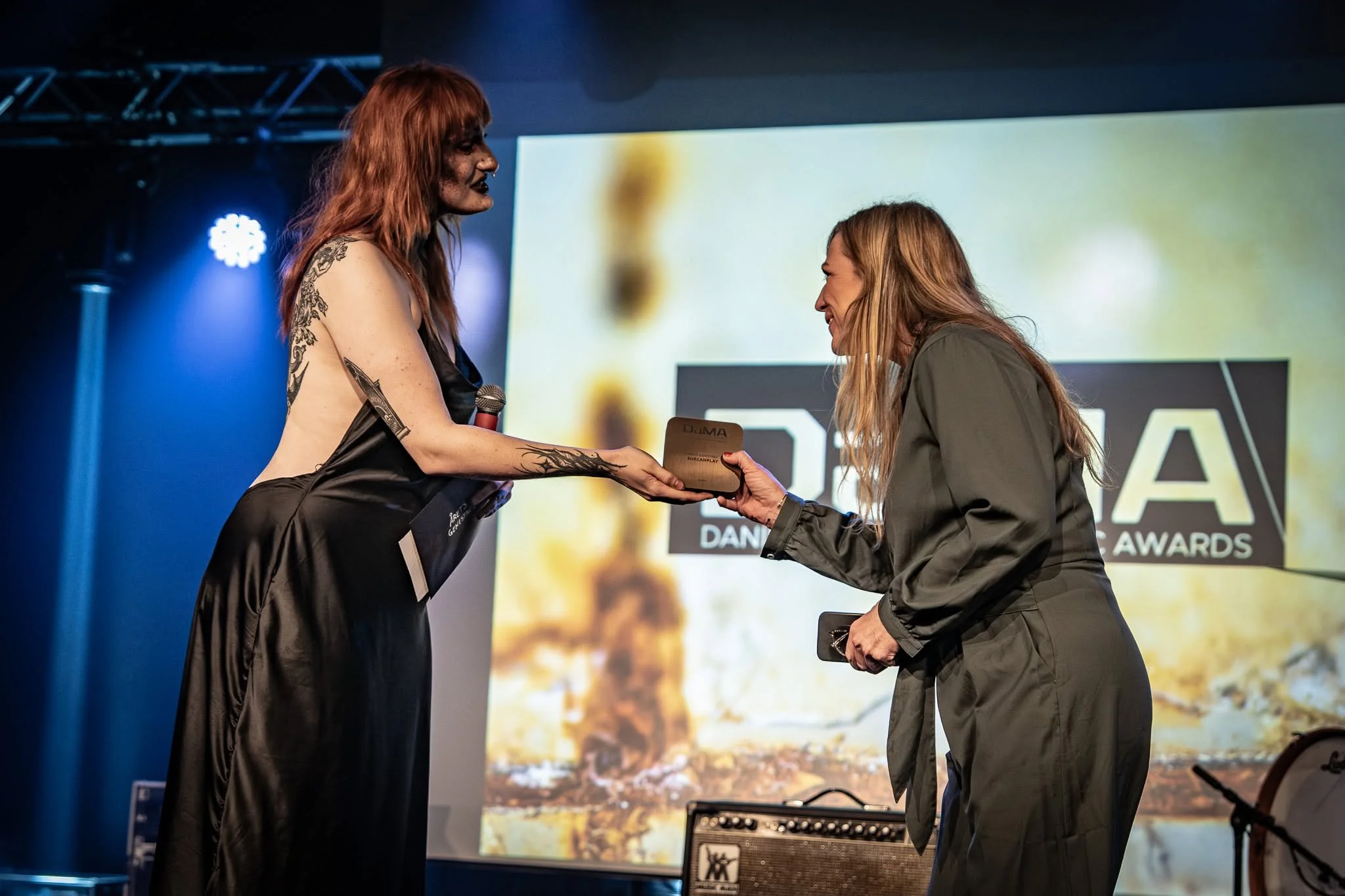 Two women are on stage during an awards ceremony. One woman with red hair and tattoos is presenting an award to the other woman with long blonde hair, who is smiling. The background displays a large screen and stage lights.