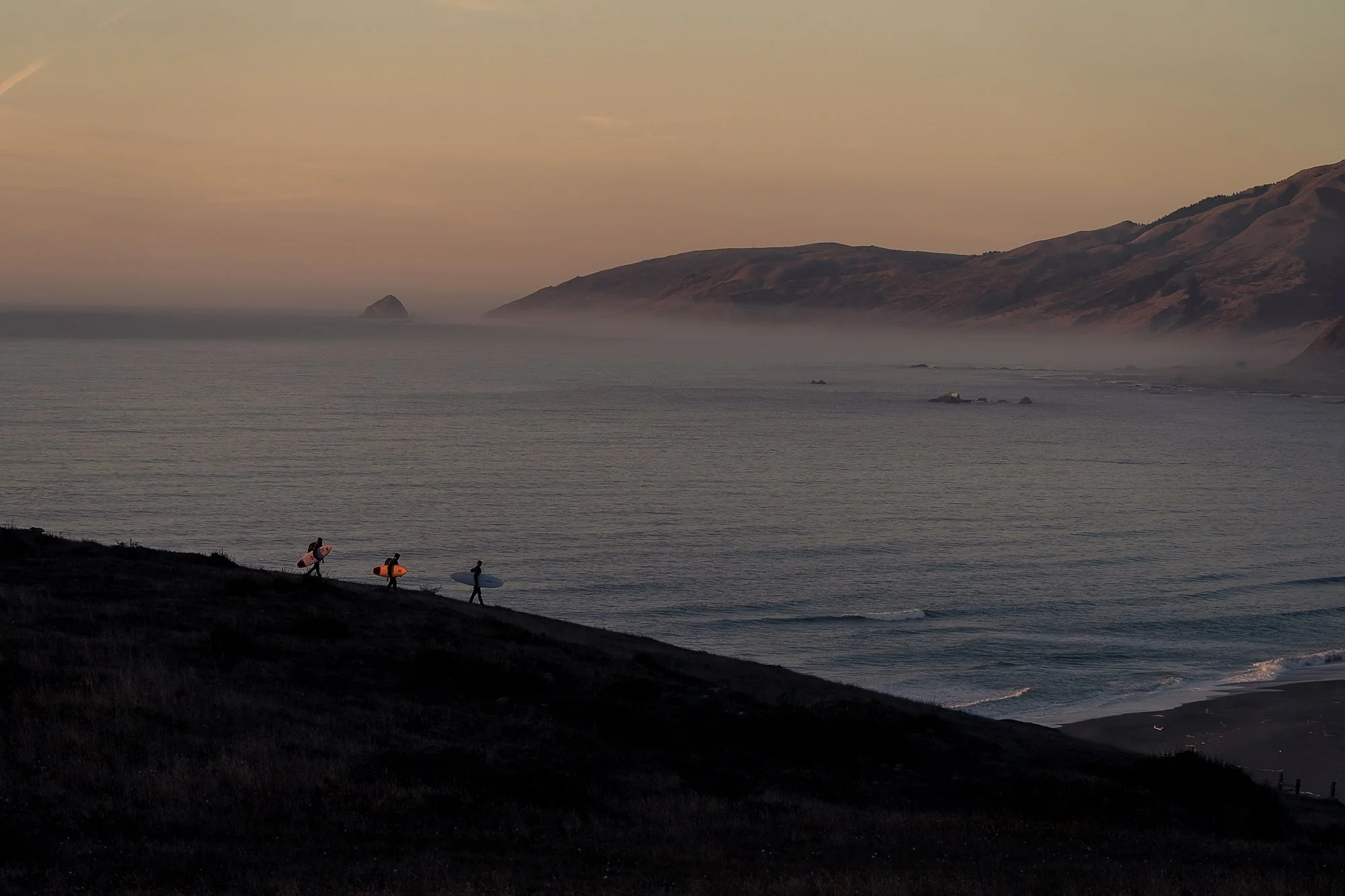 A photo by Mark McInnis of three surfers carrying boards along a ridge overlooking the ocean.