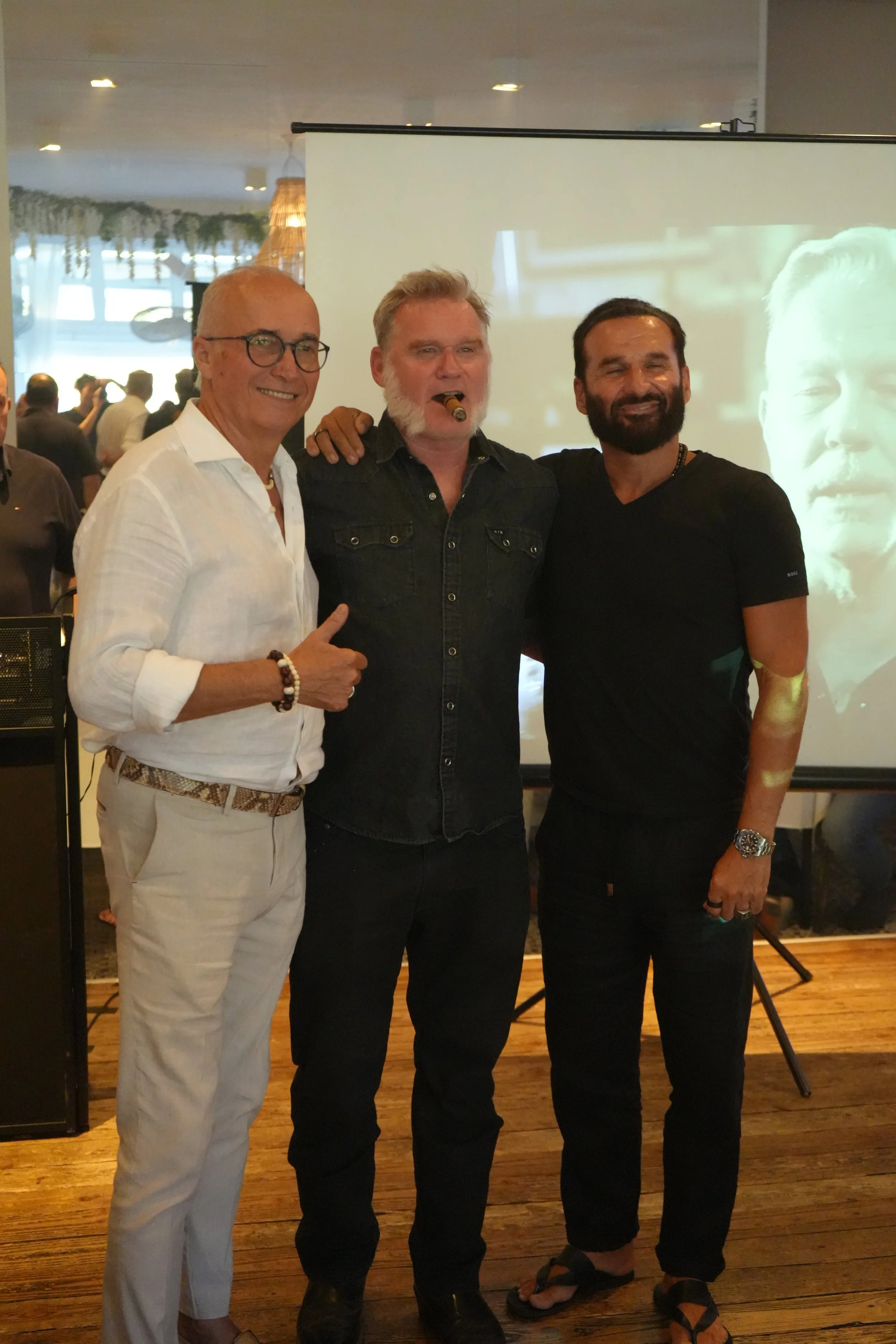 Three men standing together at an indoor event, smiling and posing for the photo. The man on the left is wearing glasses, a white shirt, and beige pants. The man in the middle has a beard, is smoking a cigar, and is wearing a dark denim shirt. The ma