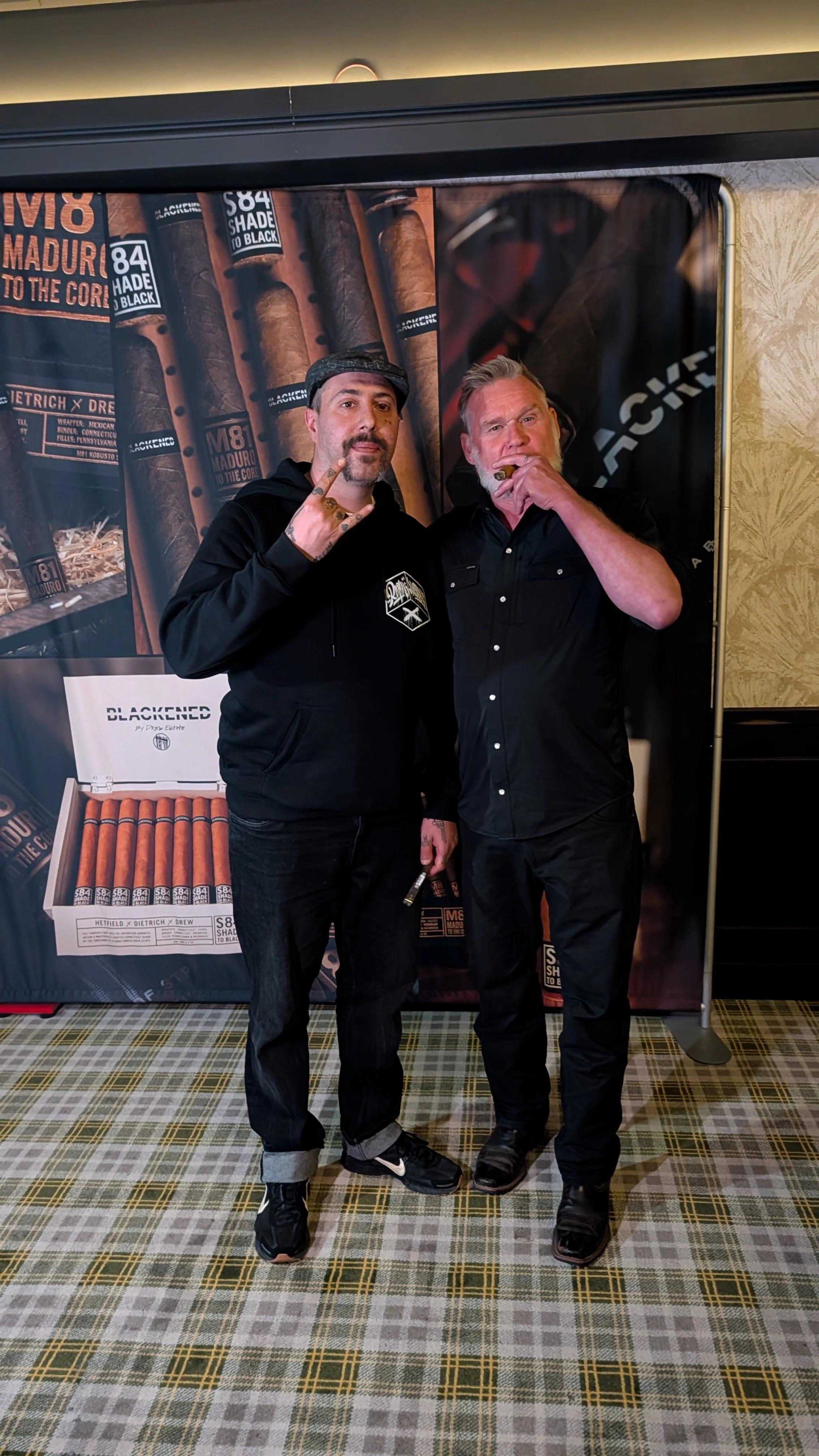 Two men standing in front of a cigar-themed backdrop at an event, one making a hand gesture and the other holding a cigar.