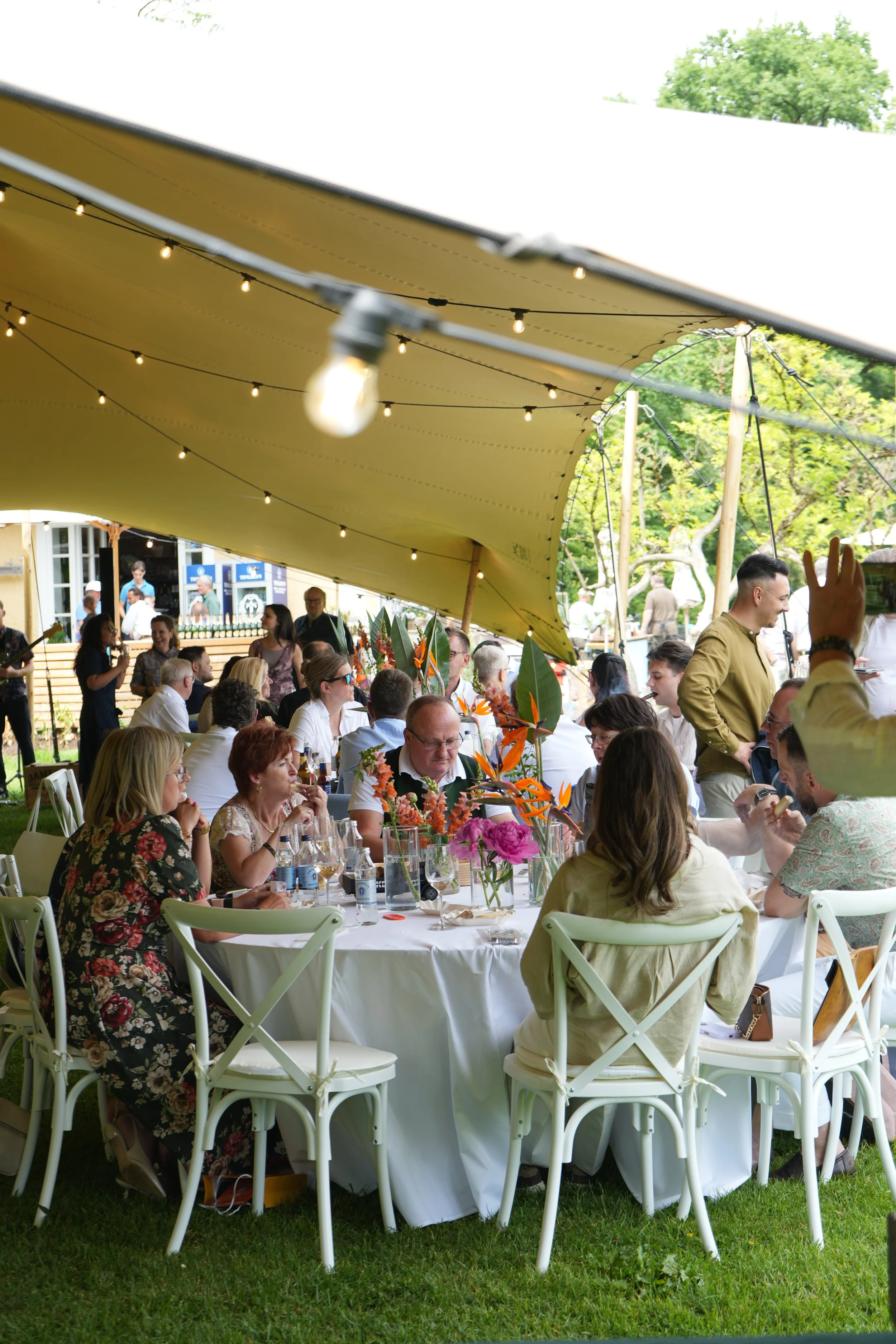 People seated at a long outdoor banquet table under a large cream-colored canopy, enjoying a gathering with floral centerpieces, surrounded by green trees and string lights.