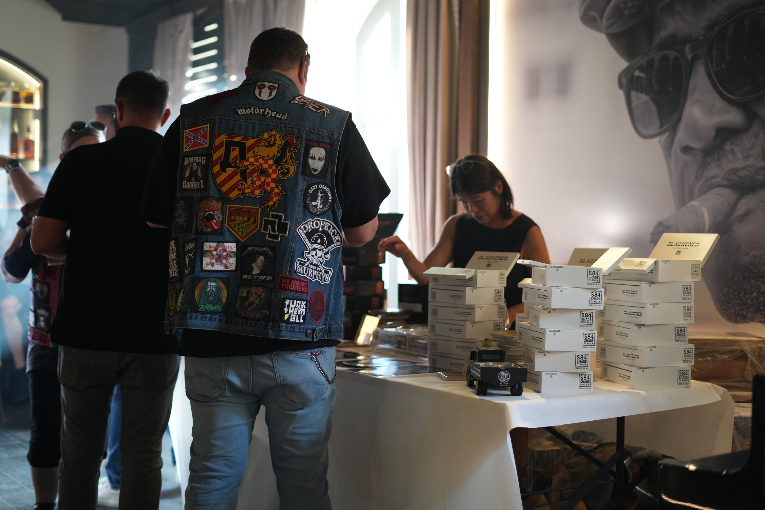 People shopping at a store counter with boxes of products on display, including skincare items labeled 'Shade to Black' and a person in a denim vest with patches and graphic designs.