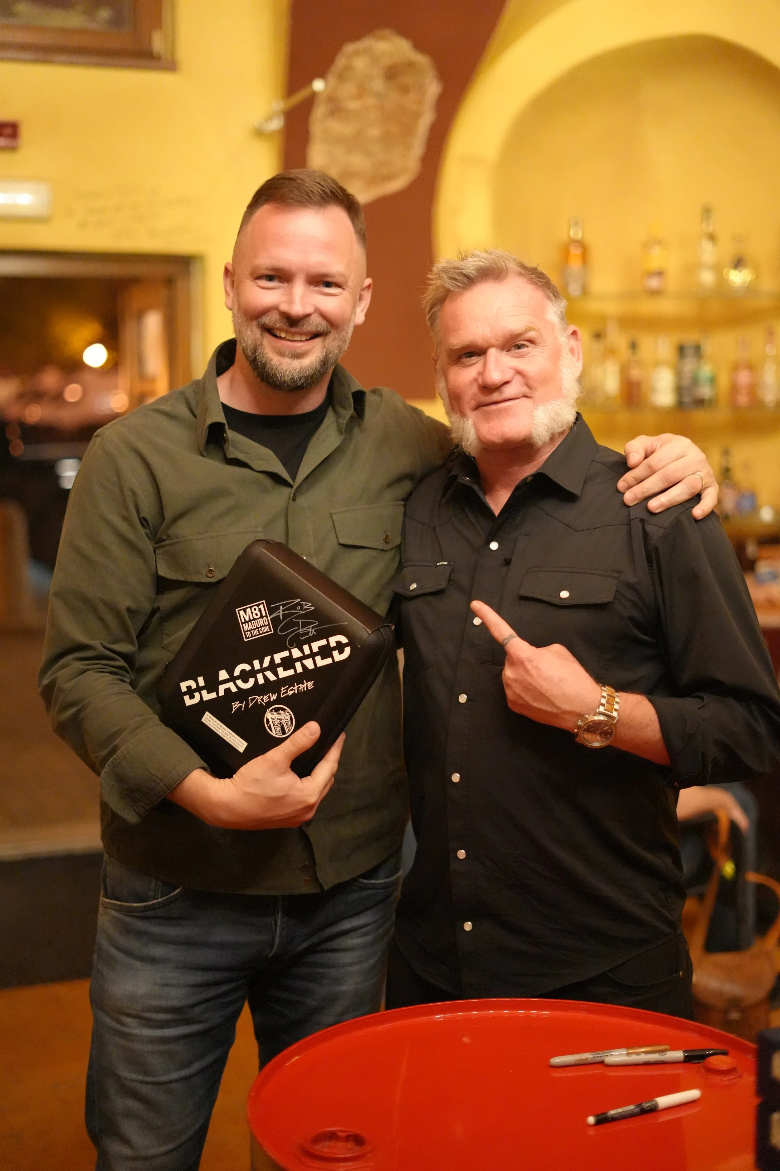 Two men are standing together in a cozy bar or restaurant, smiling at the camera. The man on the left is holding a book titled 'Blackened' and has a beard and short hair. The man on the right has gray hair and a beard, wearing a black shirt, and is p
