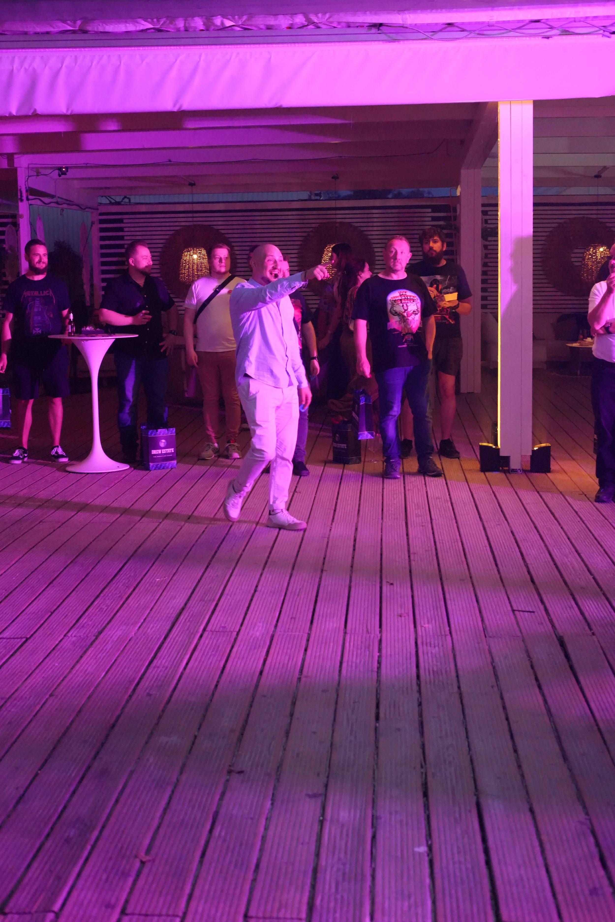 People at a party on a deck with purple lighting, some holding drinks, and one person smiling and pointing.
