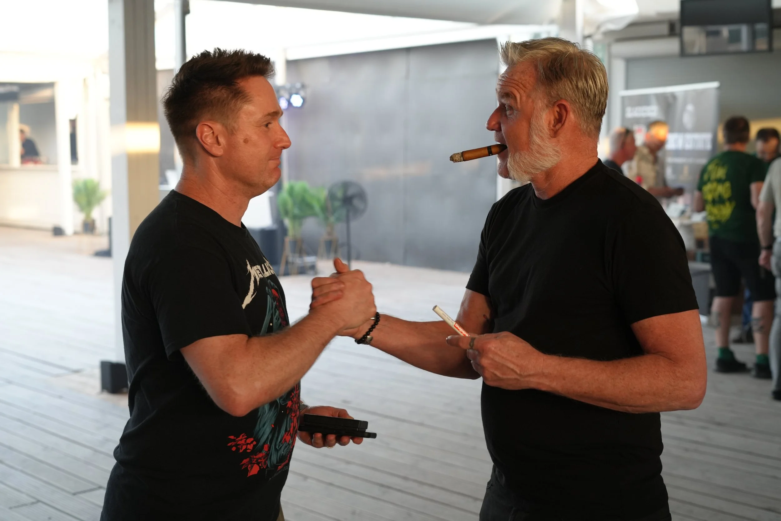 Two men are shaking hands and smiling at each other while one of them holds a cigar in his mouth, in a lively indoor setting.