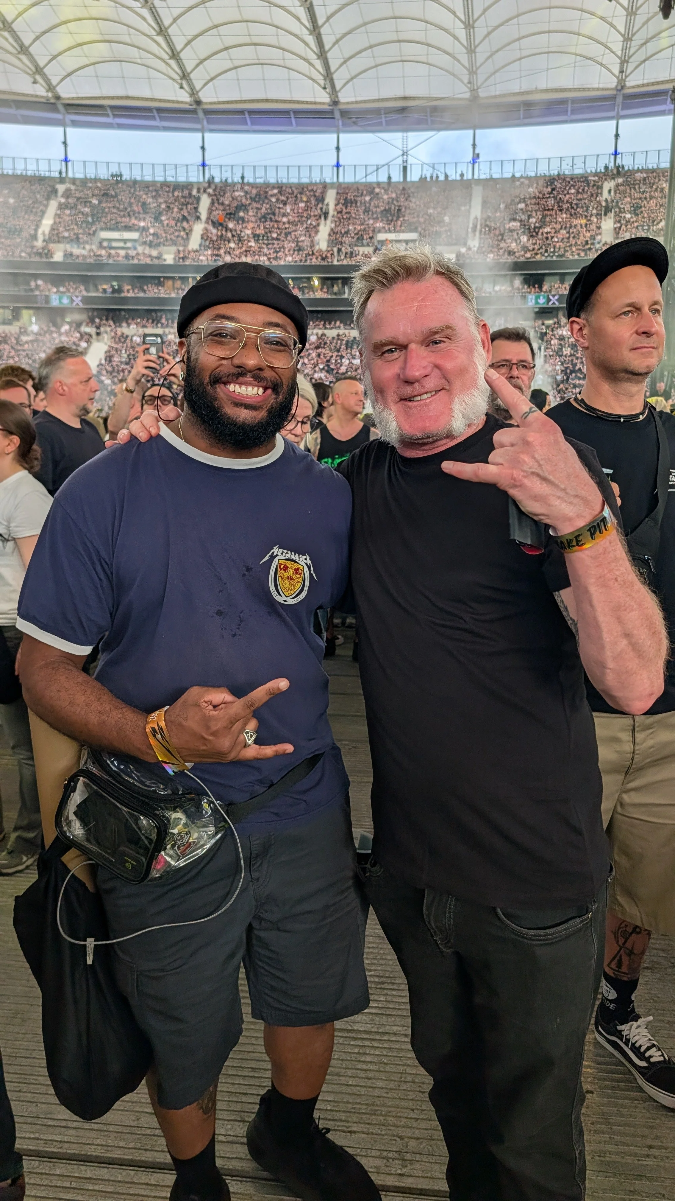 Two men posing together at a concert, one giving the rock on hand gesture, with a large crowd and stadium in the background.