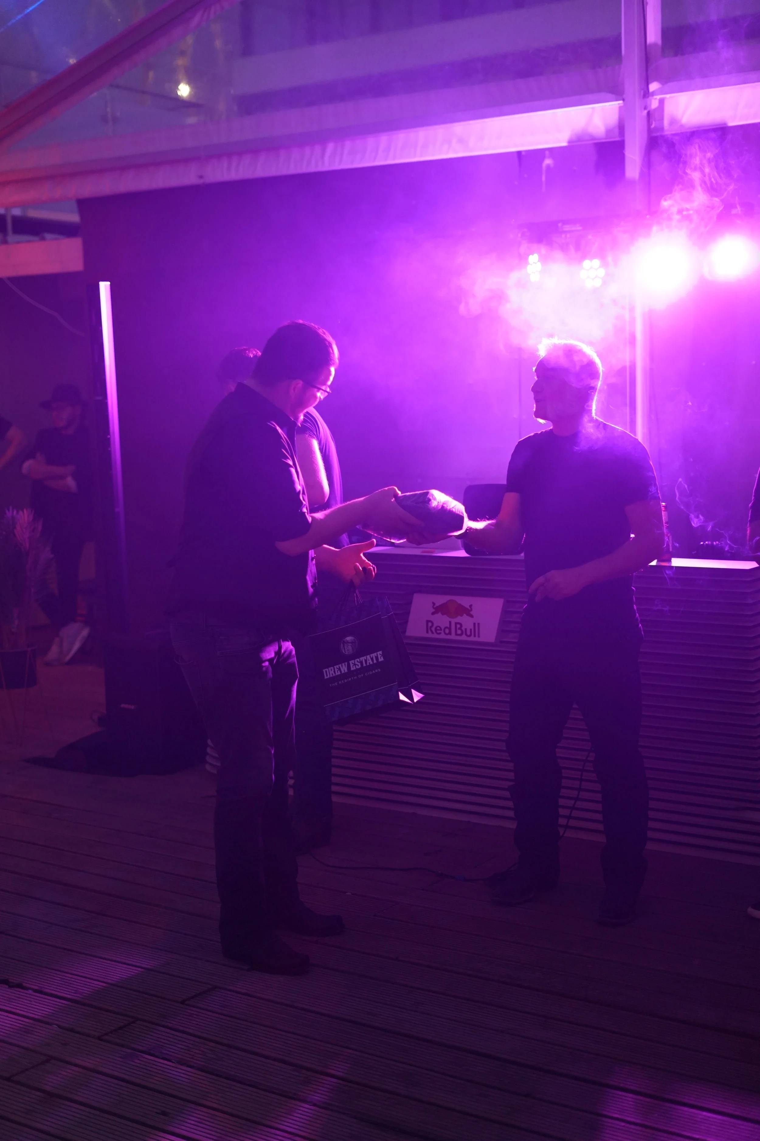 Two men exchanging a handshake or an item at a party or event with purple stage lighting. There is a Red Bull paper bag and a sign for Drew Estate on the counter. People are in the background, some observing and others listening or talking.