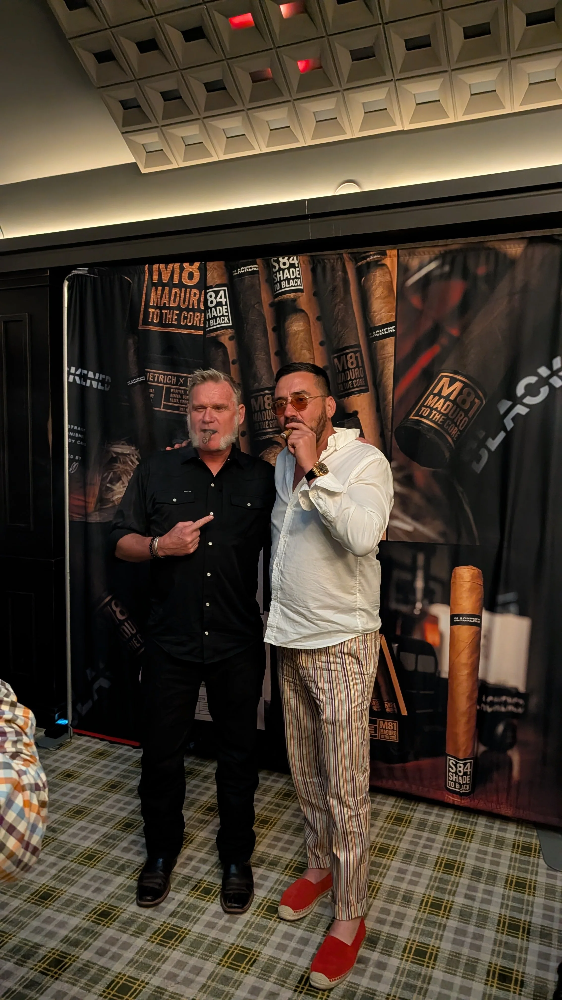 Two men standing in front of a cigar-themed backdrop, one with white hair and beard wearing black shirt, the other with dark hair and beard wearing a white shirt and striped pants, both smoking cigars.