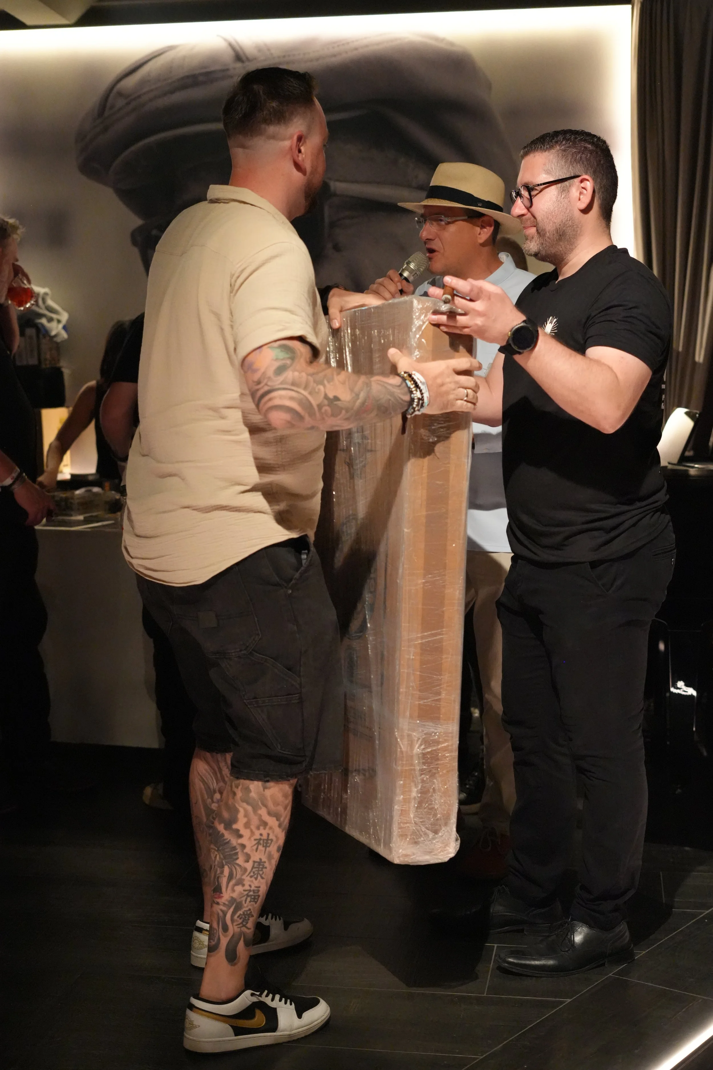Three men are gathered around a wooden table in a dimly lit room. One man with glasses and a black T-shirt listens carefully. Another man with a hat and glasses speaks into a microphone, while the third man, with tattoos and dressed casually, interac