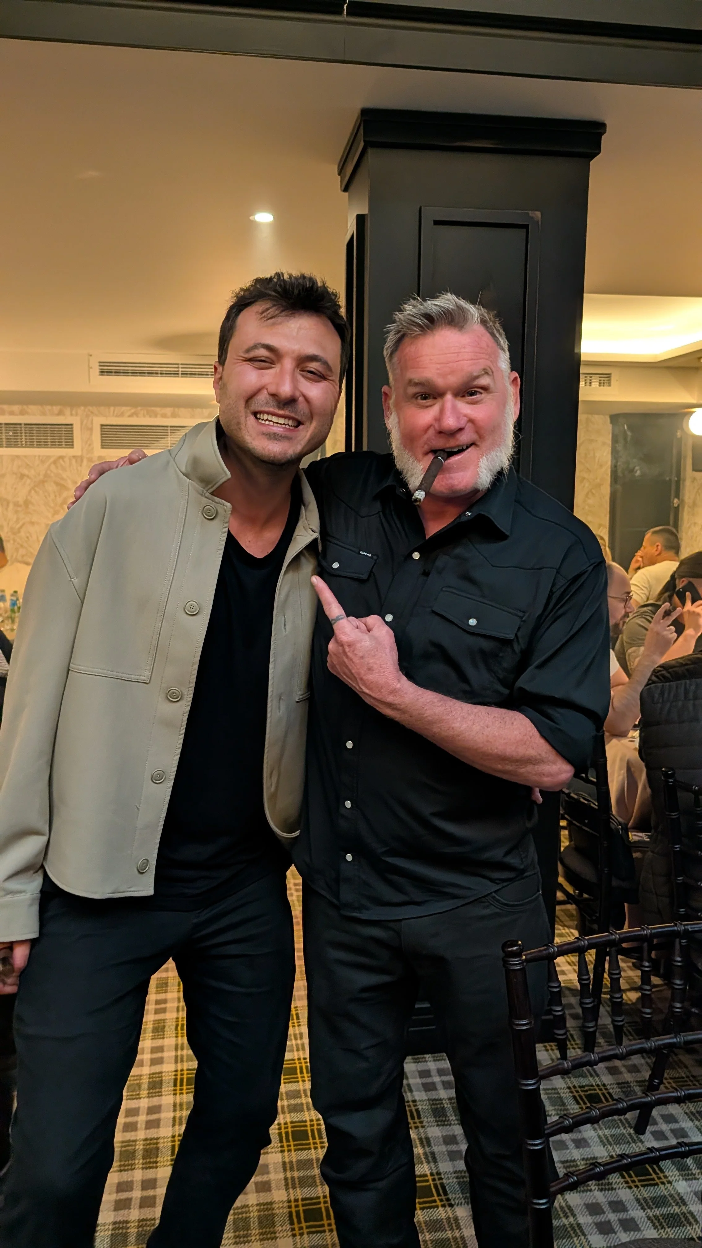 Two men smiling and posing for a photo in a restaurant, one with a cigar and pointing at the other.