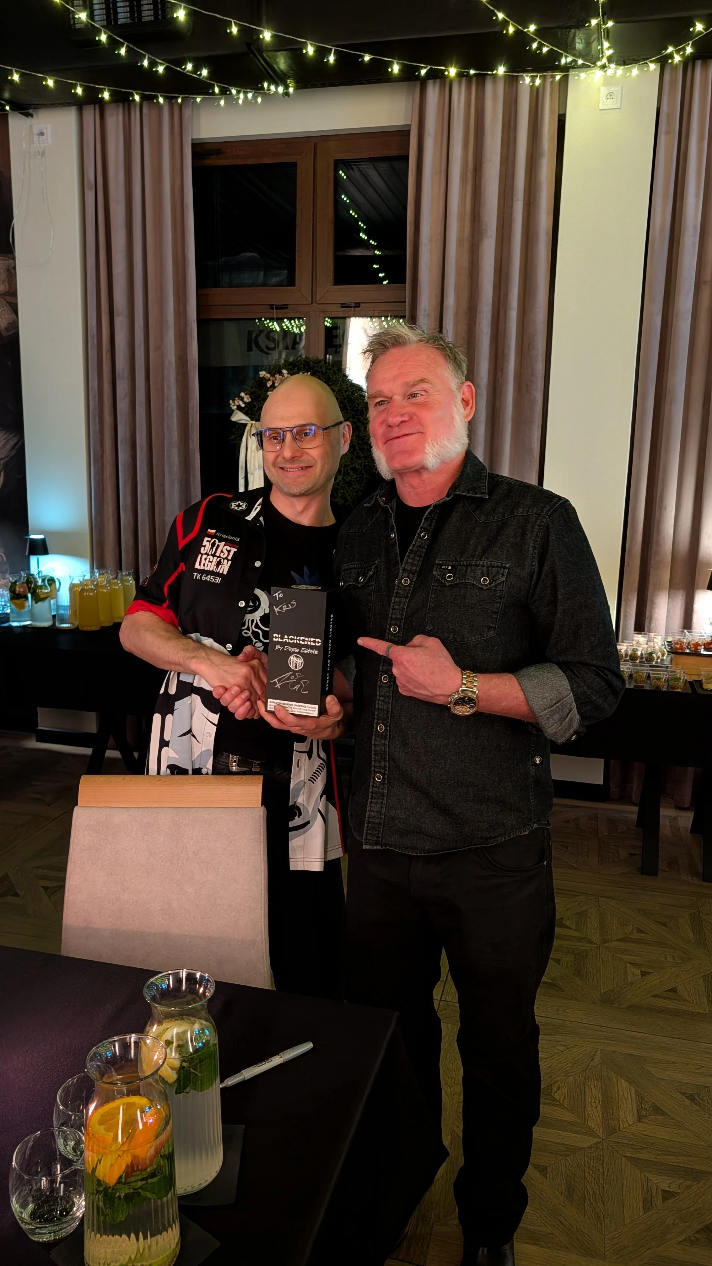 Two men are standing together in a room decorated with string lights. One man is holding a boxed item and both are smiling at the camera. There are drinks on the table in front of them and curtains in the background.