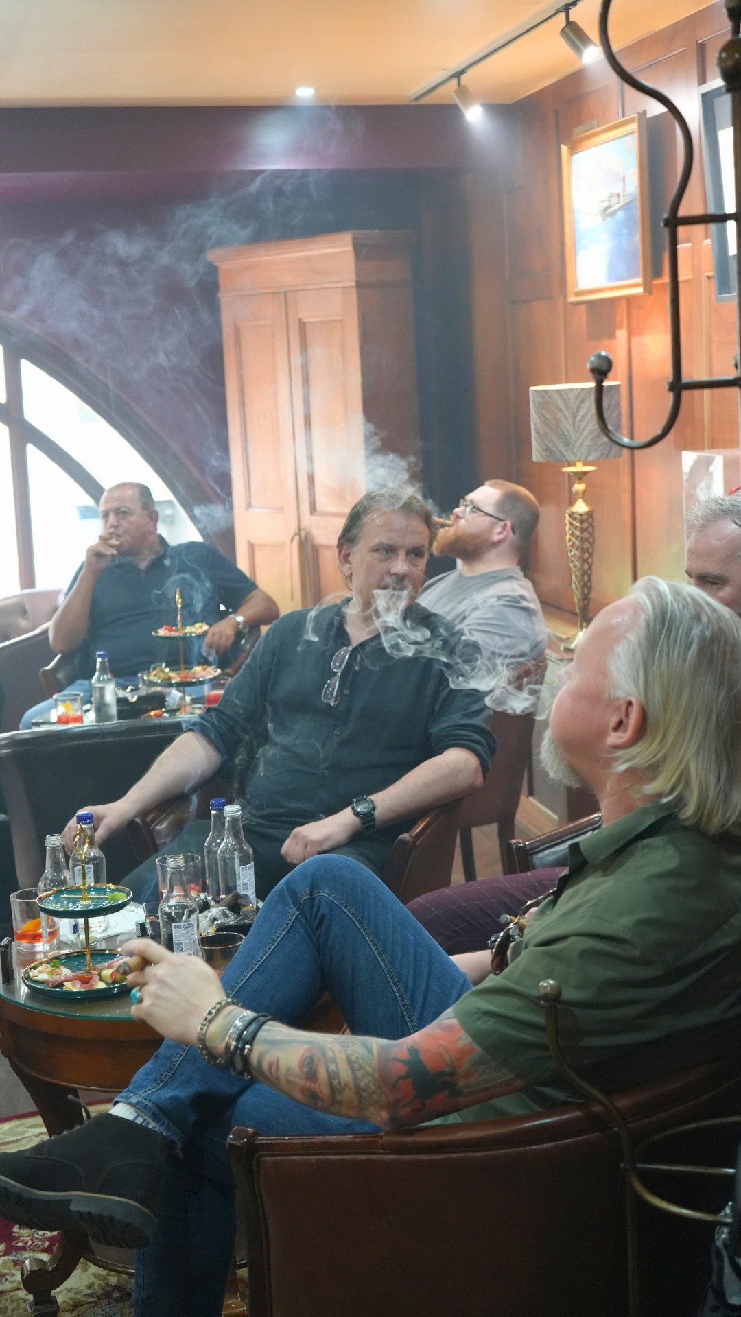 A group of men sitting in a cozy, wood-paneled room with paintings on the wall, smoking and drinking, with tables of drinks and snacks in front of them.
