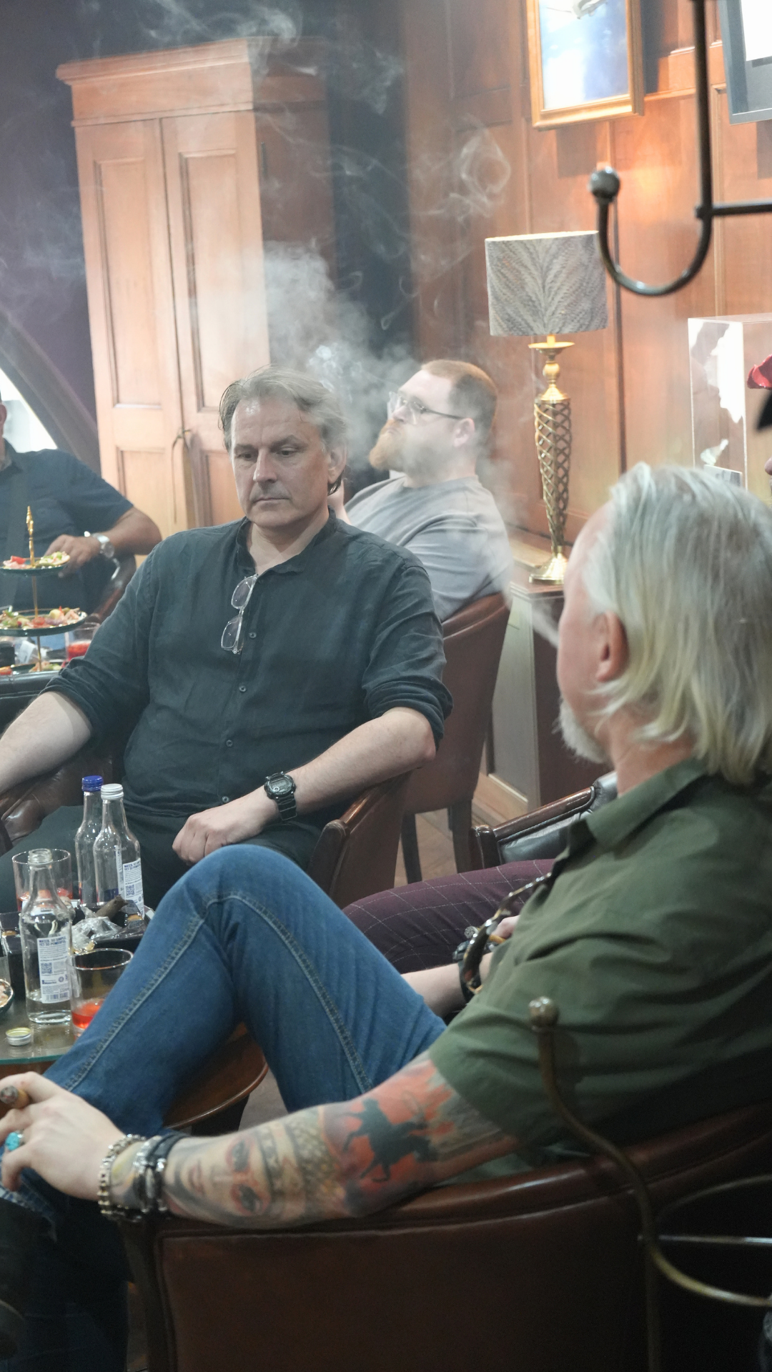 People sitting in a room with wooden paneling, smoke in the air, and a table with drinks and snacks.