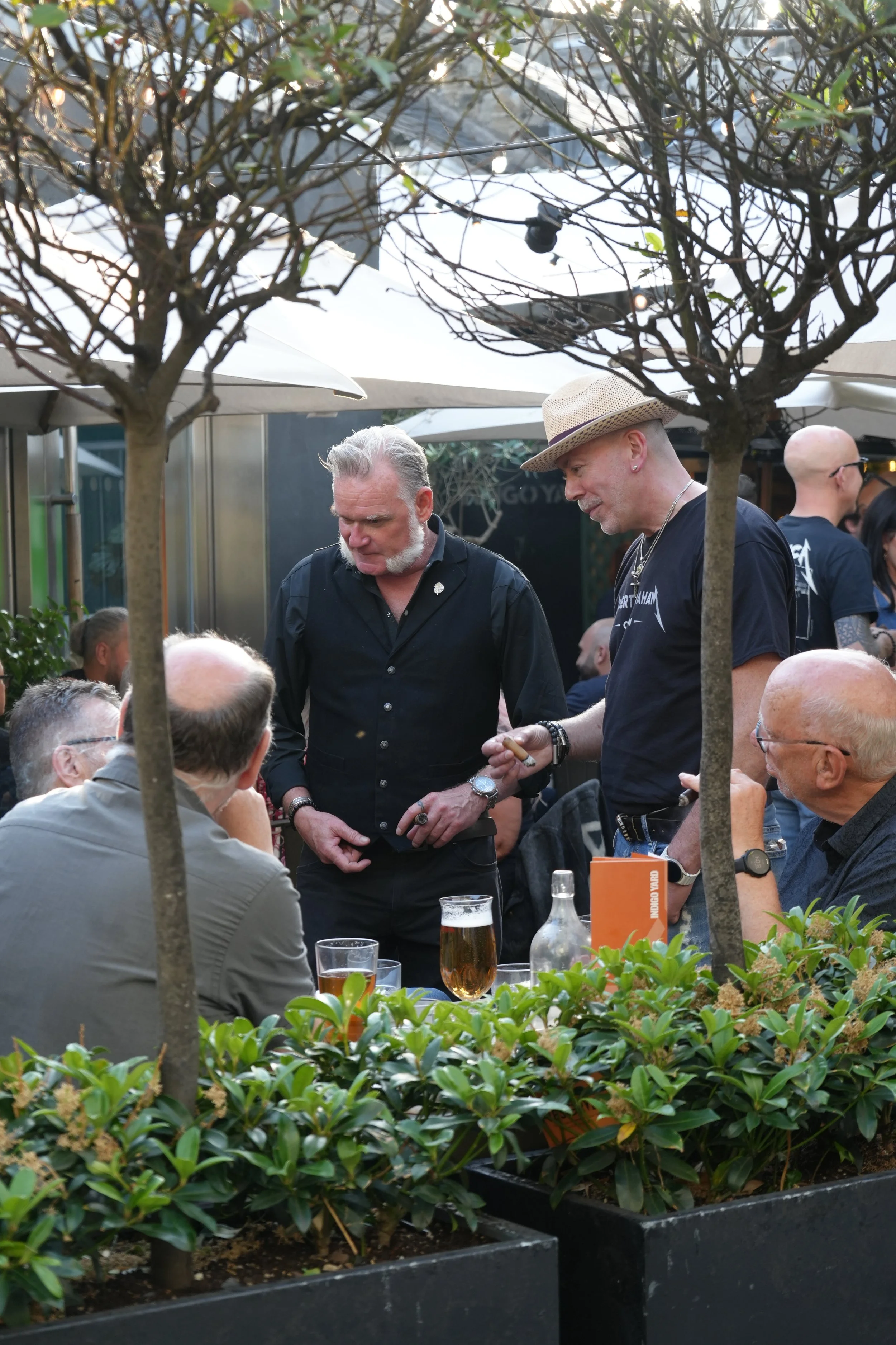 People socializing outdoors at a restaurant or bar, with two men standing, one with a straw hat and one with gray hair, flanked by others sitting at a table with drinks, surrounded by potted plants and trees.