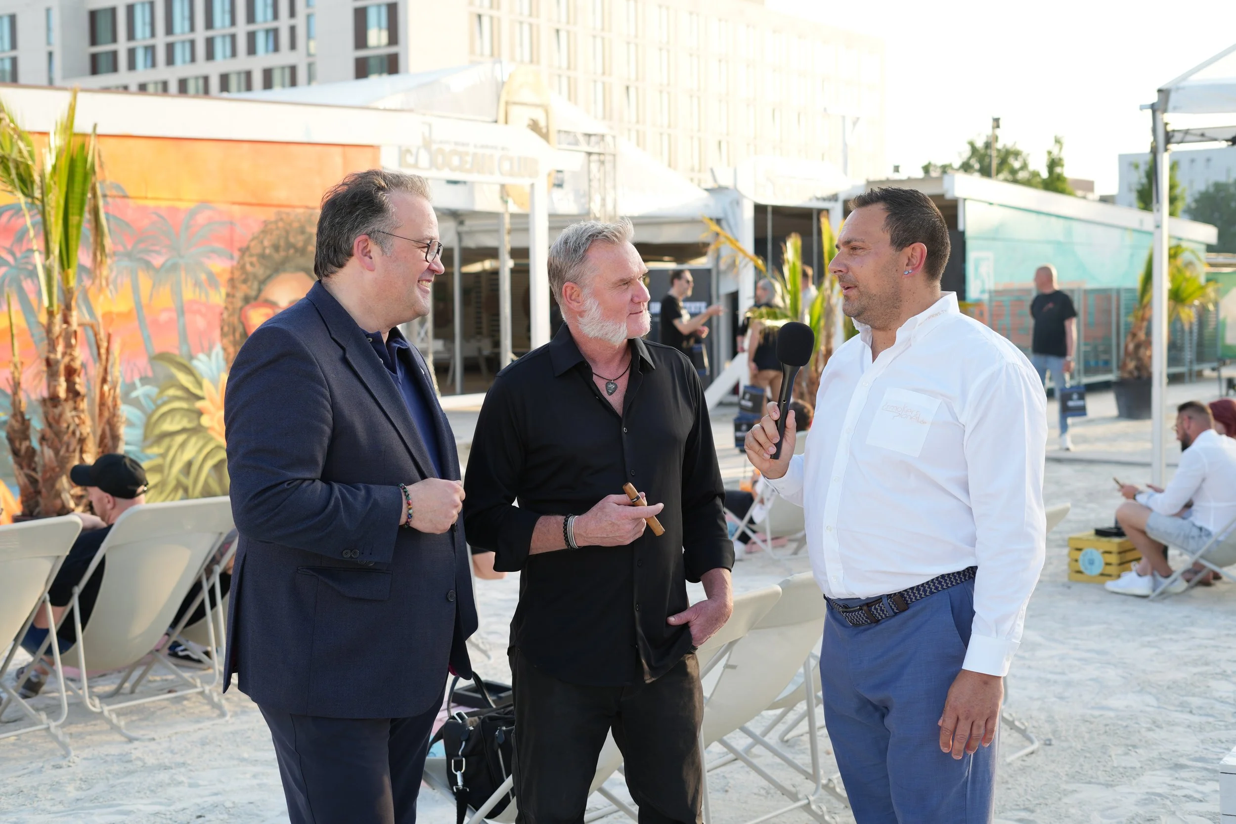 Three men engaging in conversation on a beach during daytime, with seated people and buildings in the background.