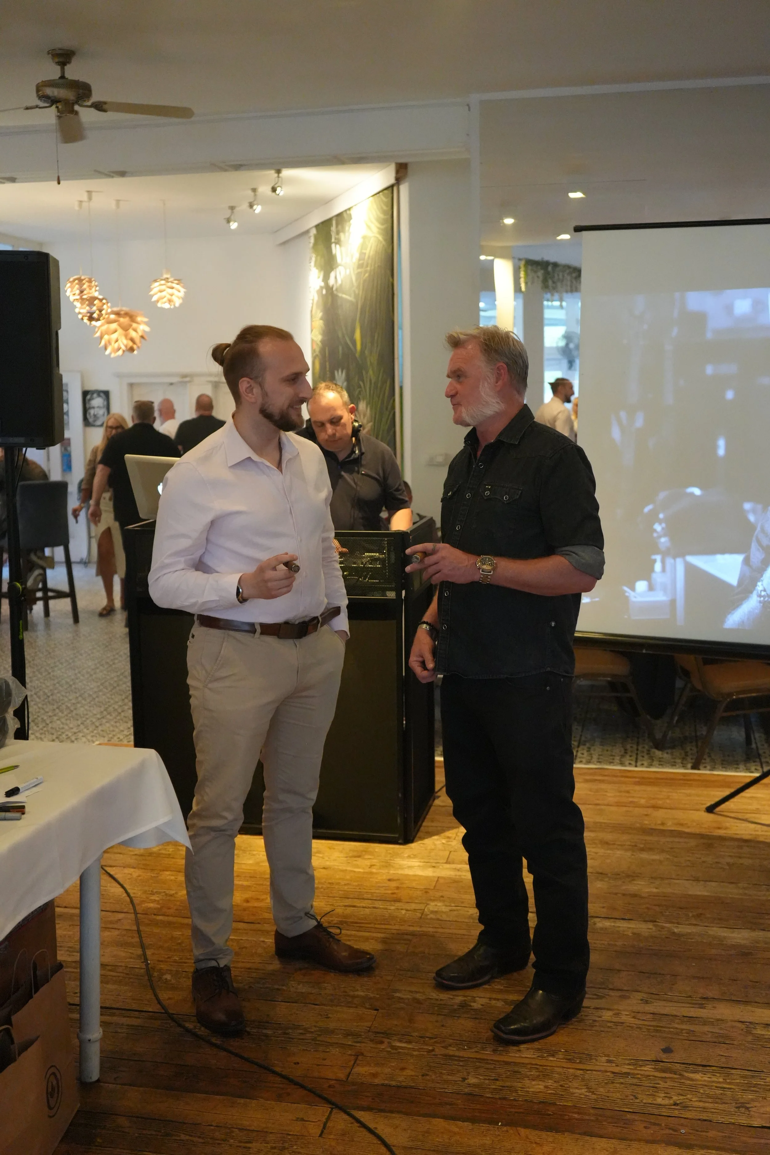 Two men are talking in a conference or social event setting, with one wearing a white shirt and beige pants and the other in a black shirt and black pants. Several people are in the background near a laptop, with decorative lighting and artwork on th