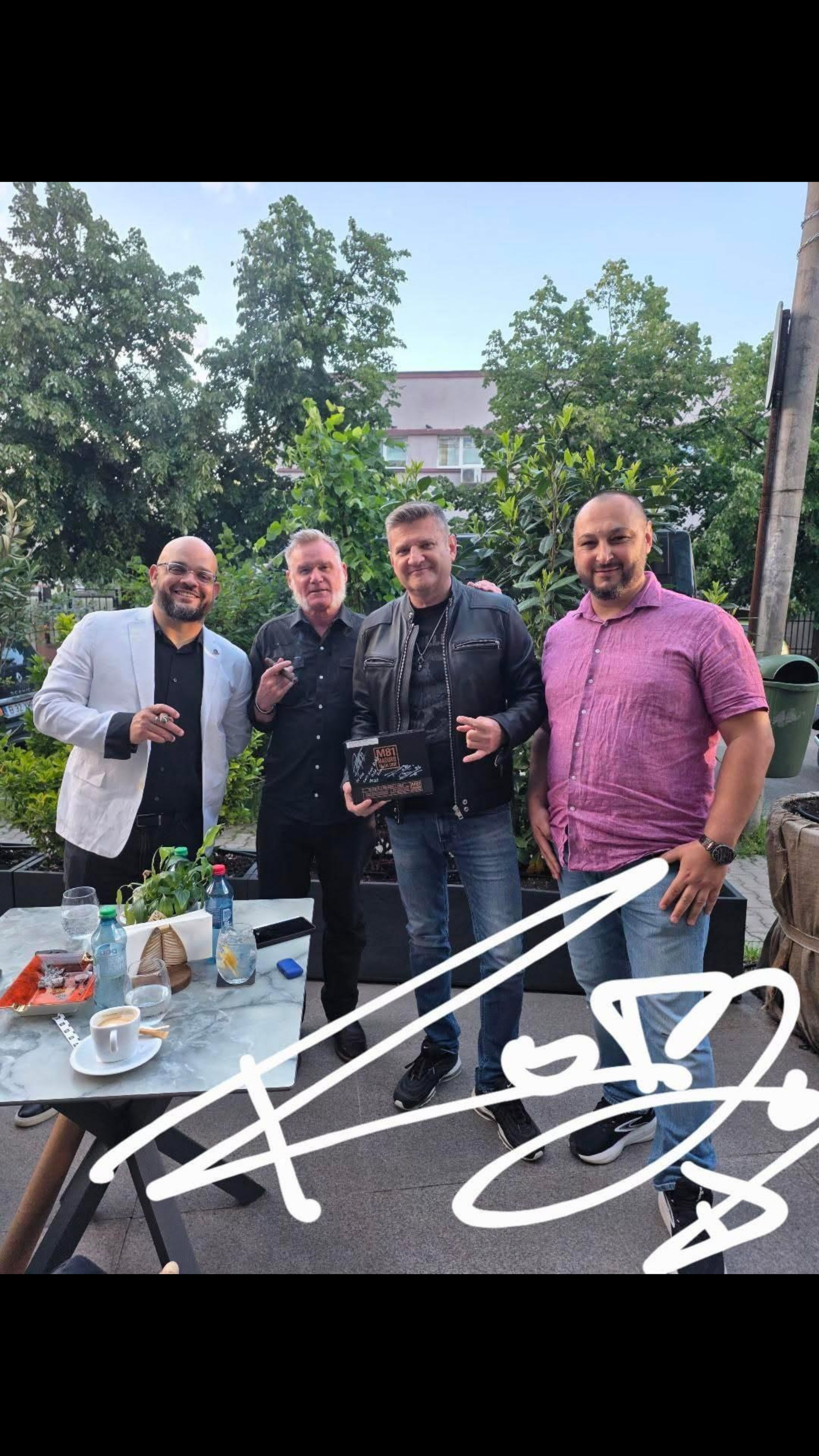 Four men standing outdoors in front of greenery, smiling at the camera, with a table of drinks and snacks in front of them.