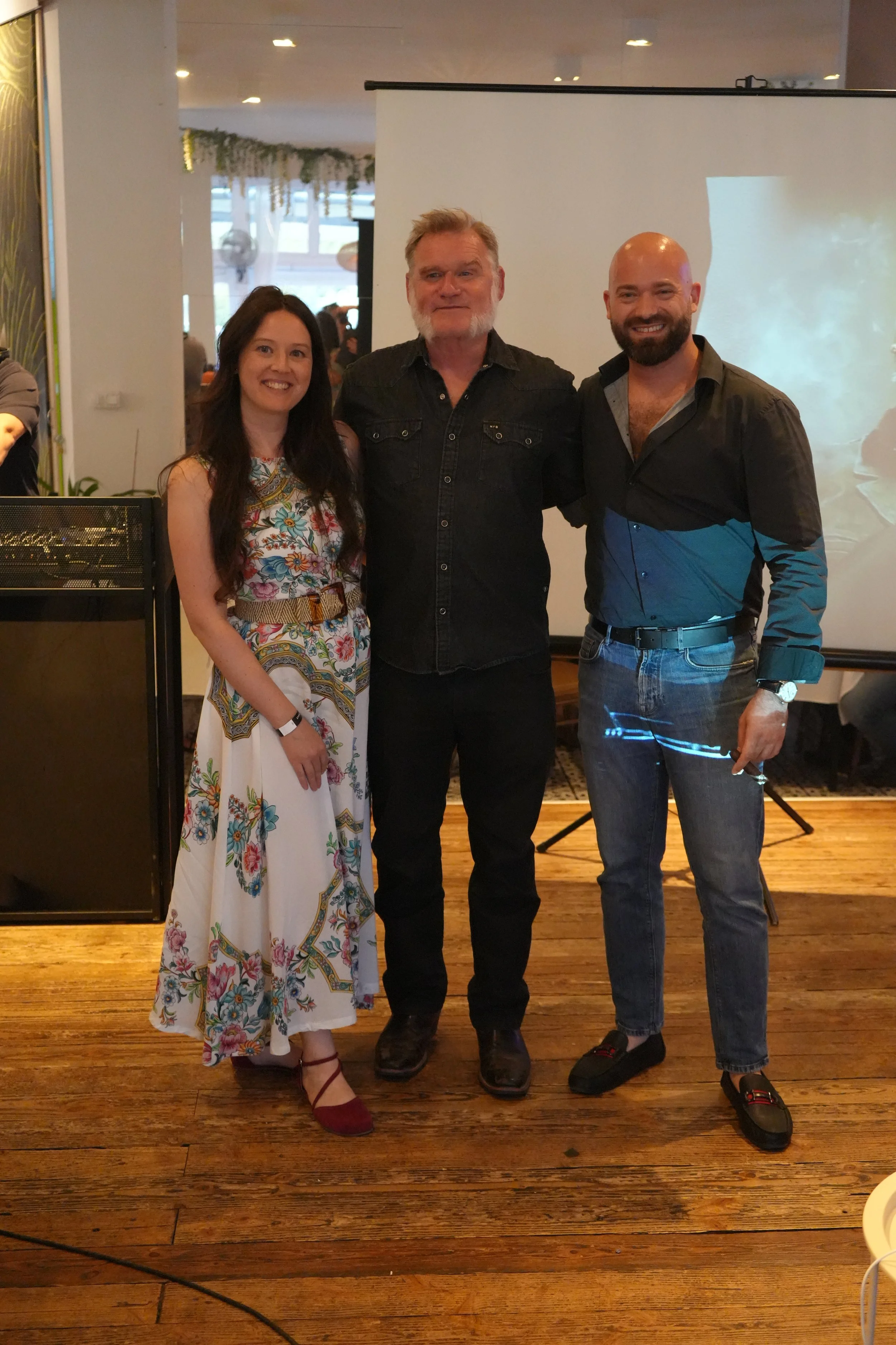 Three people standing together in a room with a wooden floor. On the left, a woman in a floral dress with a belt and maroon shoes smiling. In the middle, a man with a beard and mustache wearing a black denim shirt and black trousers. On the right, a 