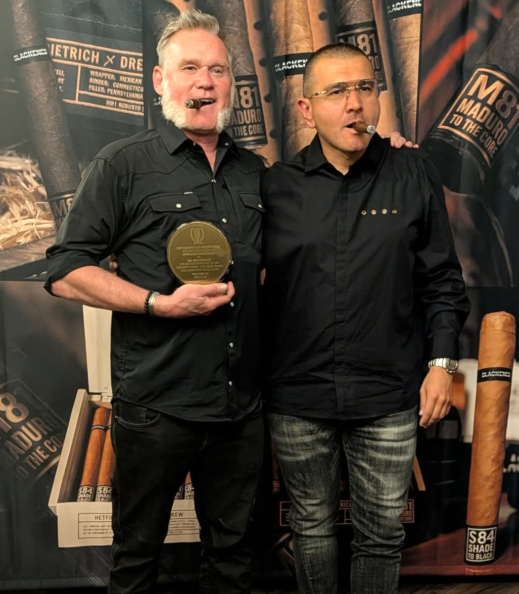 Two men standing together with cigars and one holding a trophy, in front of a backdrop with cigar branding.