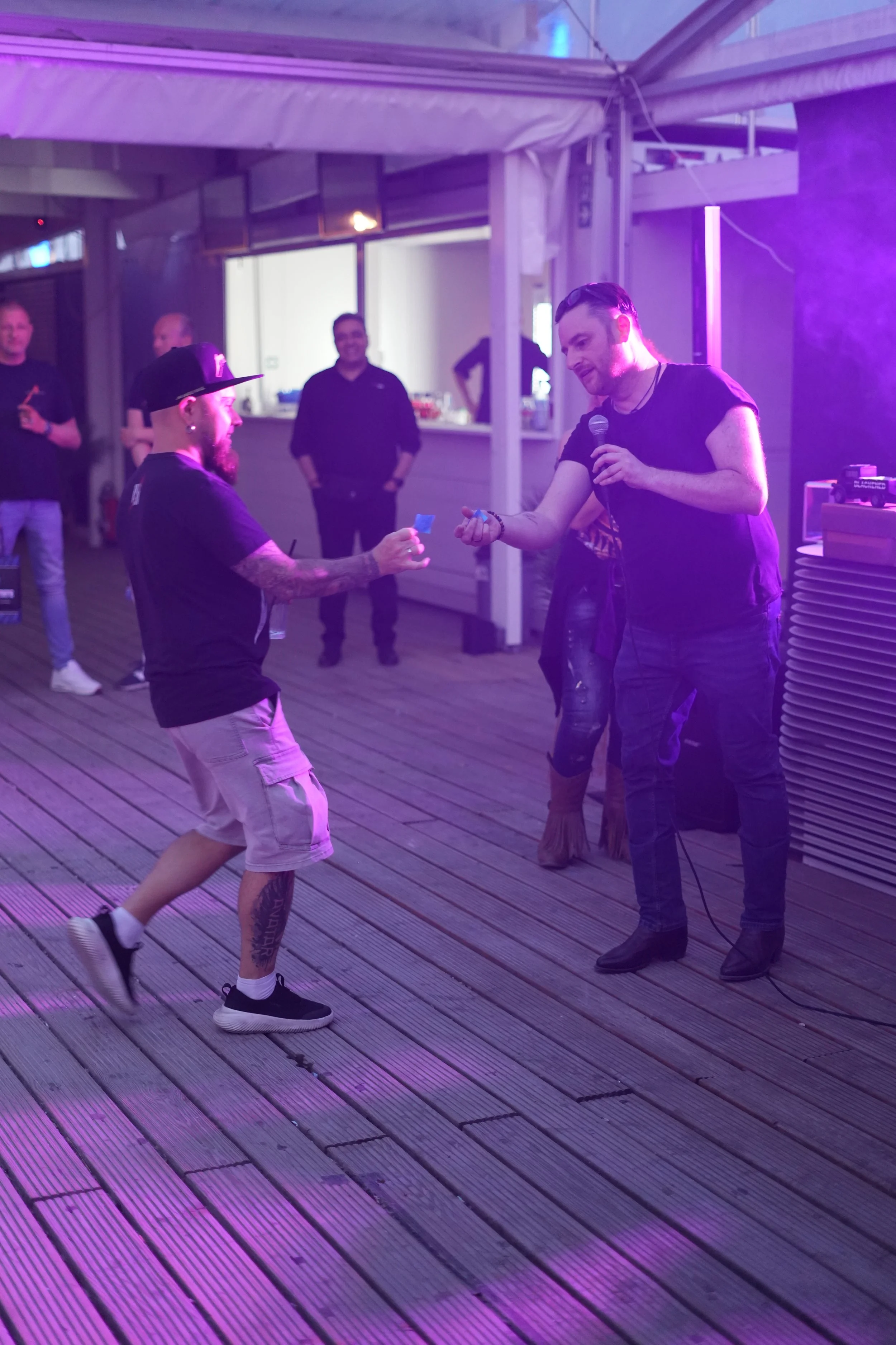 A man with a beard and tattooed arms wearing a black t-shirt, baseball cap, and shorts engaged in a dance or energetic movement with another man holding a microphone, dressed in black with leather pants, on a wooden deck with purple lighting. Several