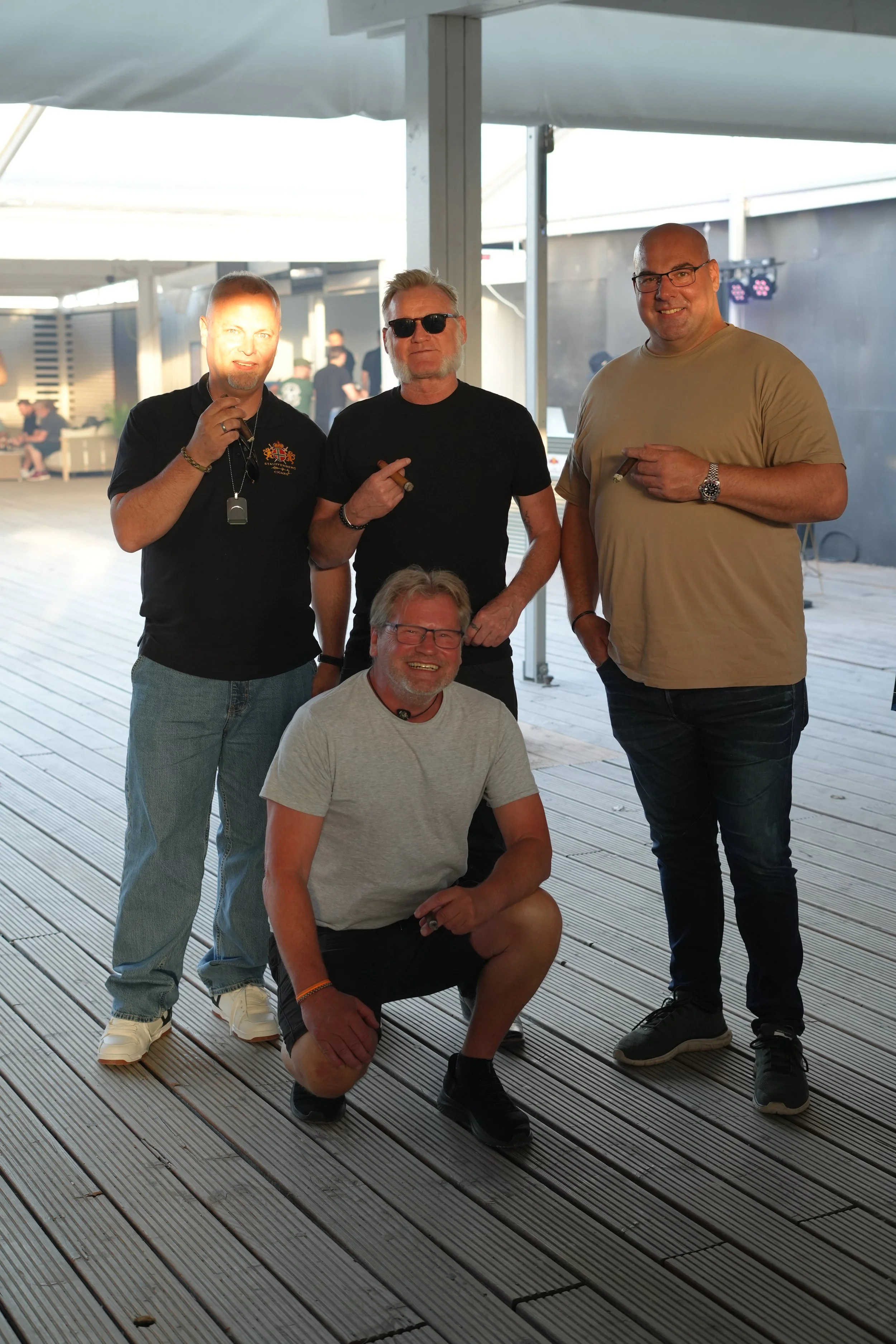 Four men posing for a photo on a wooden deck, with some in casual clothing and sunglasses, smiling and holding cigars.